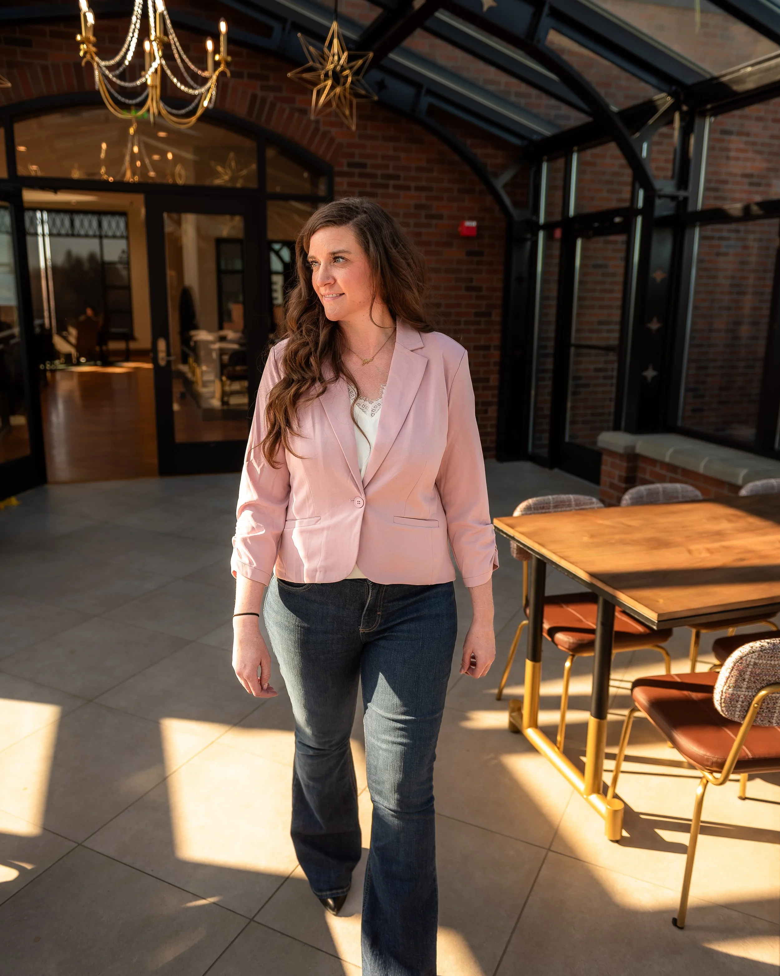 A woman with long brown hair wearing a light pink blazer, white top, and dark jeans walking inside a sunlit indoor space with brick walls and glass panels.