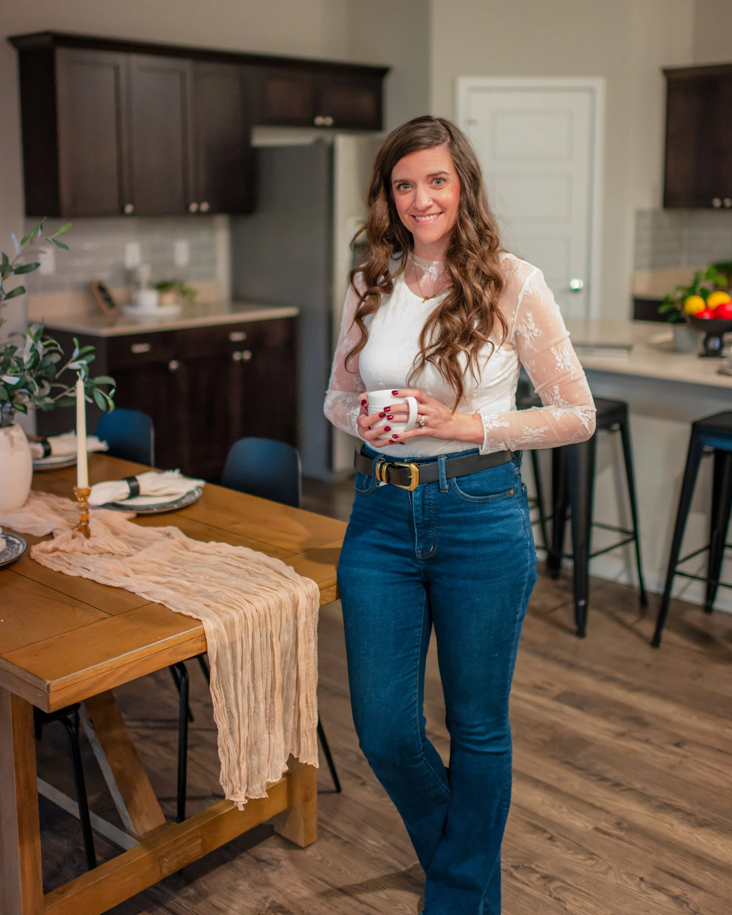 A woman with long, wavy brown hair wearing a white lace long-sleeve top and blue jeans, holding a white mug, standing in a modern kitchen with dark wooden cabinets, a dining table with a beige cloth, and a bowl of lemons on the counter.