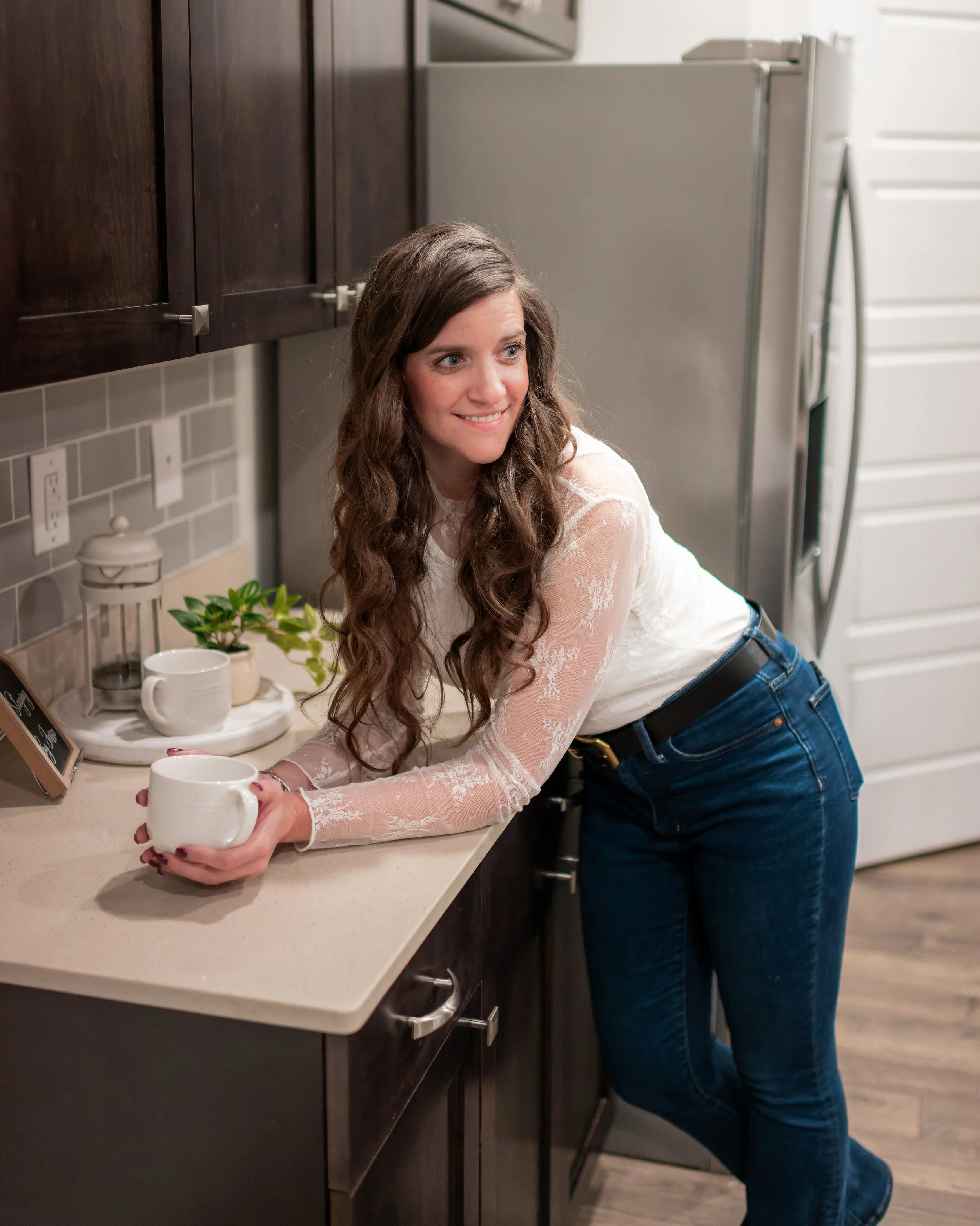 A woman with long, wavy brown hair leaning on a kitchen countertop, smiling, holding a white coffee mug, with a refrigerator and dark wood cabinets behind her.