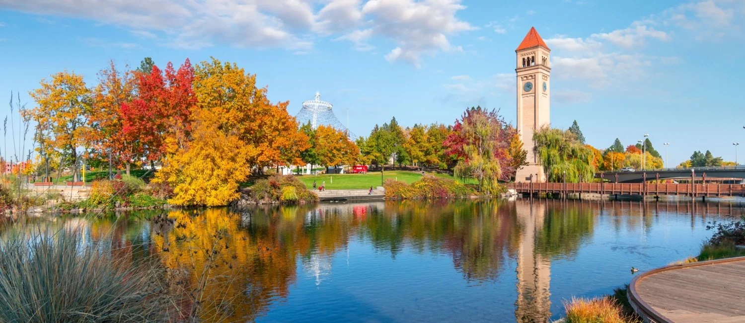 A scenic view of a river with colorful autumn trees, a tall clock tower, and a bright blue sky with clouds.