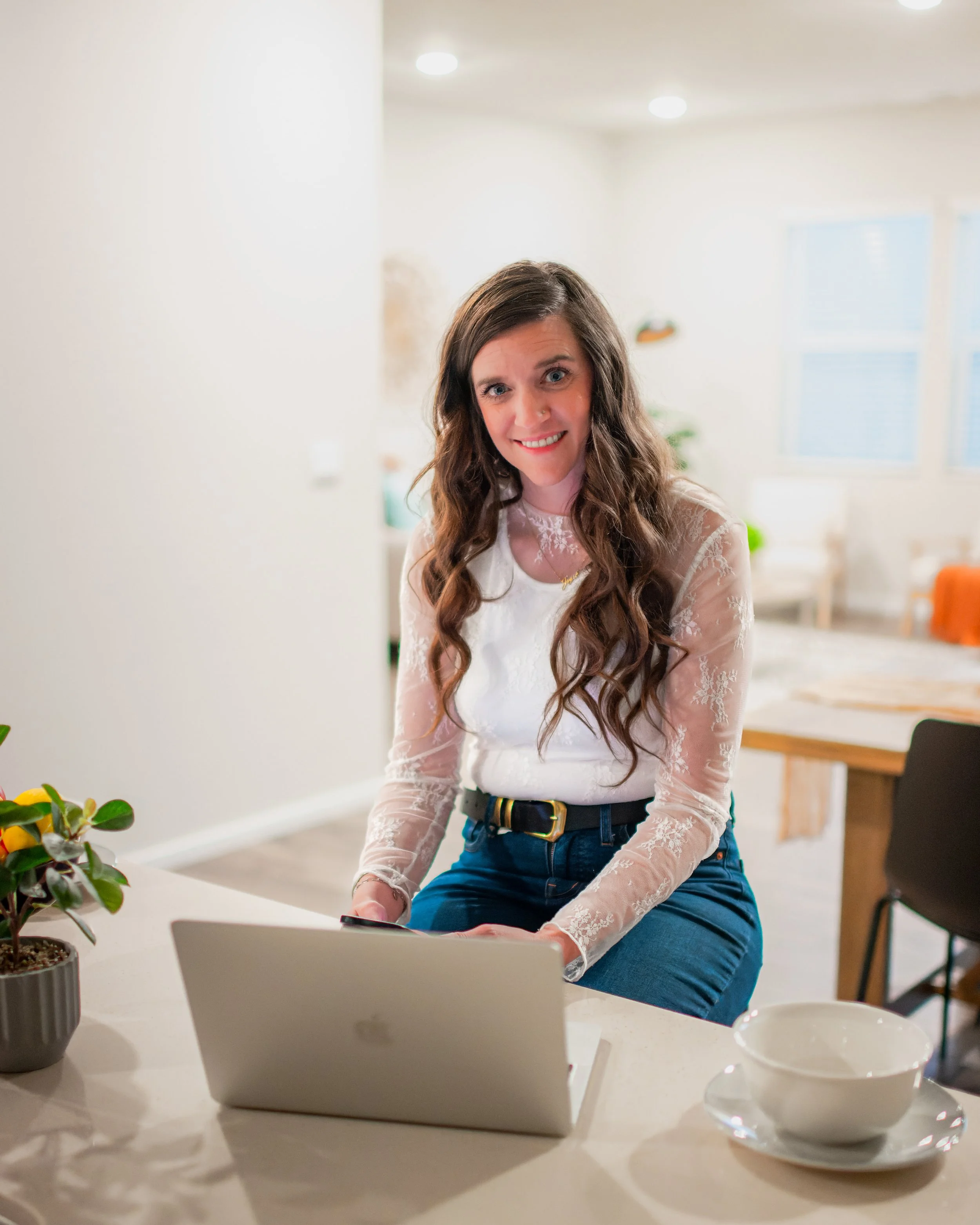A woman with long, wavy brown hair is sitting at a kitchen counter, smiling at the camera. She is wearing a white lace long-sleeve top and blue jeans. There is a laptop, a small potted plant, a bowl, and a smartphone on the counter. The background shows a well-lit, modern kitchen with windows and dining area.