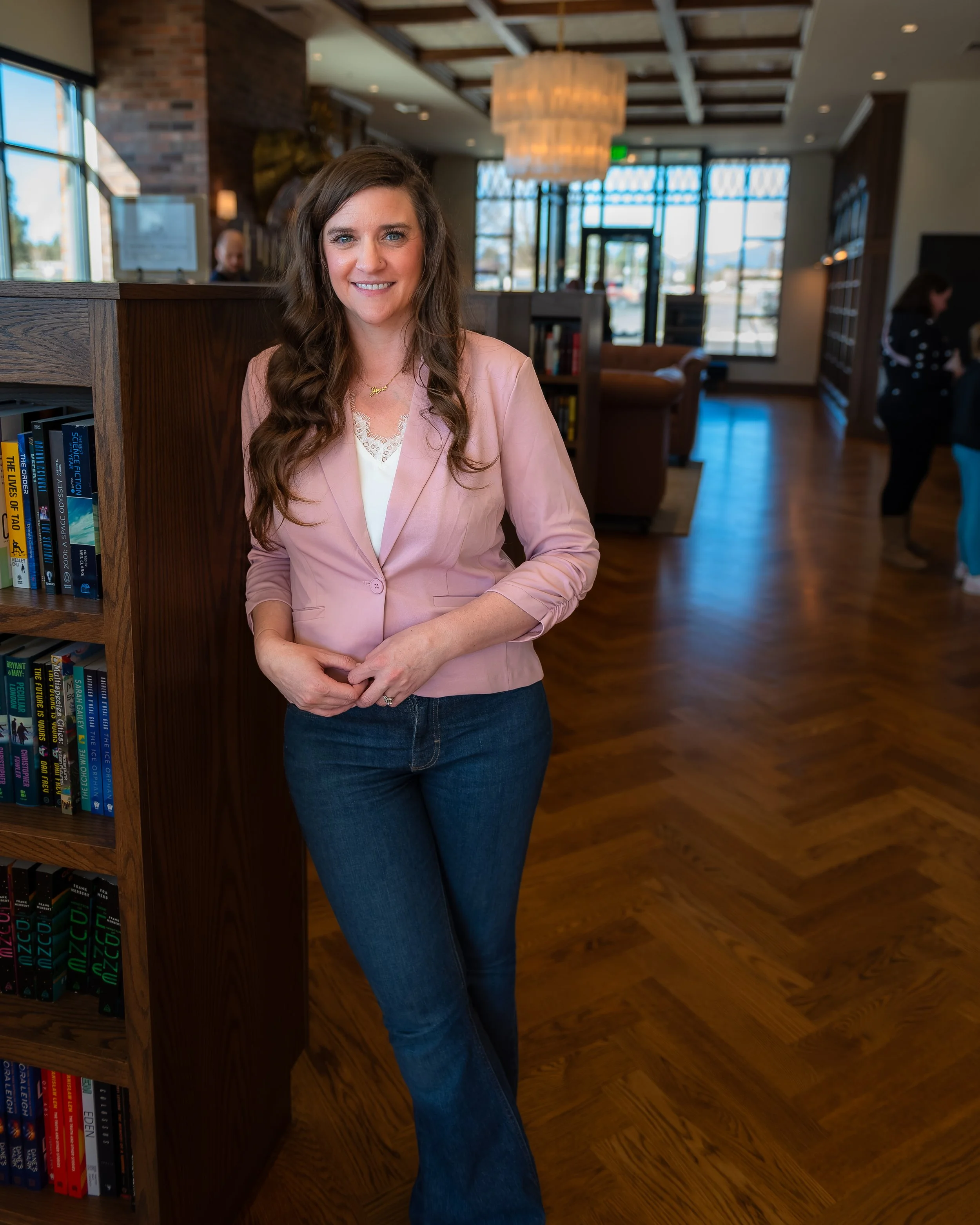 A woman with long wavy brown hair, wearing a light pink blazer, white top, and blue jeans, standing in a bookstore or library with wooden floors, large windows, and people in the background.