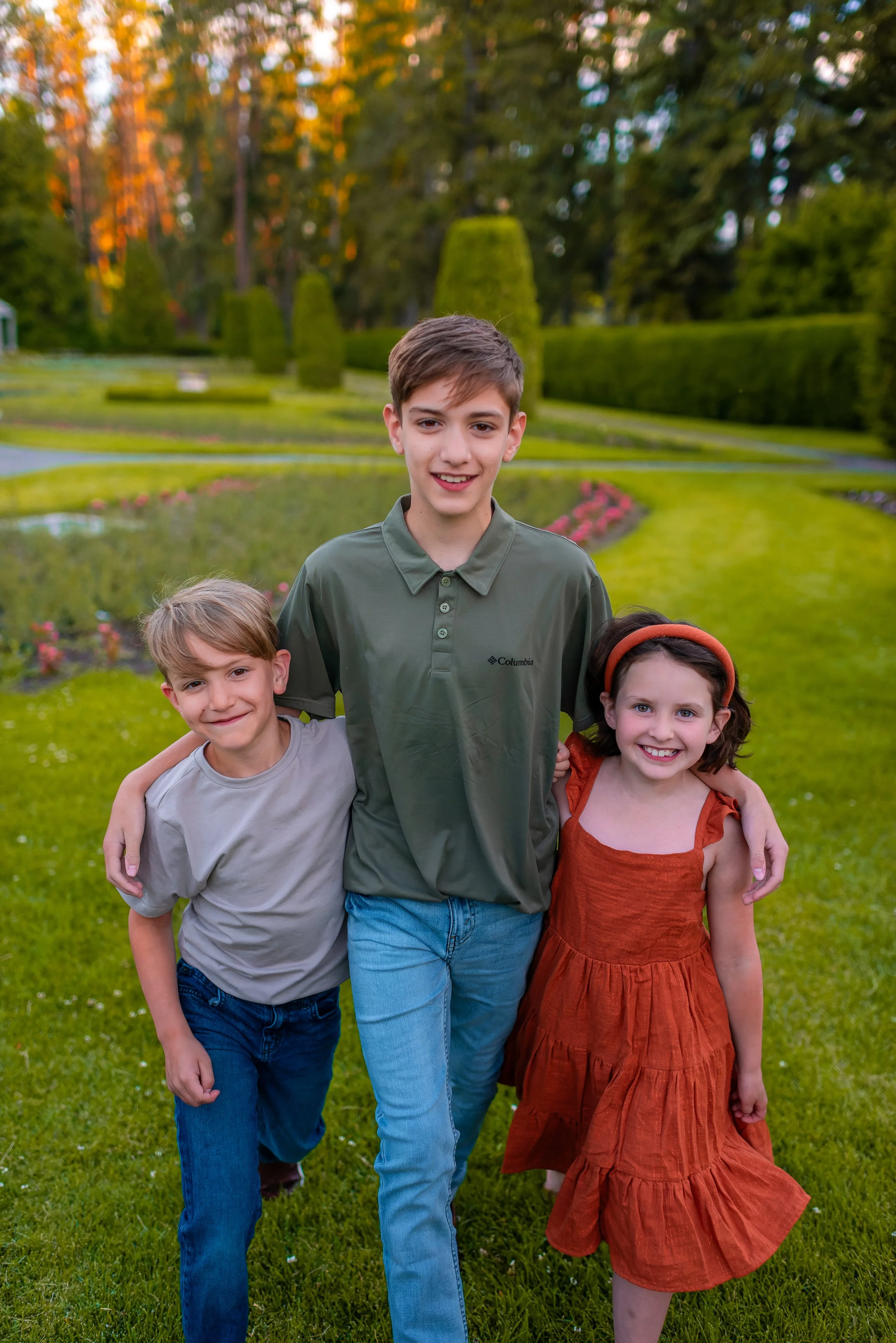 Three children, two boys and a girl, standing outside in a garden with green grass, trees, and shrubs, smiling and with their arms around each other.