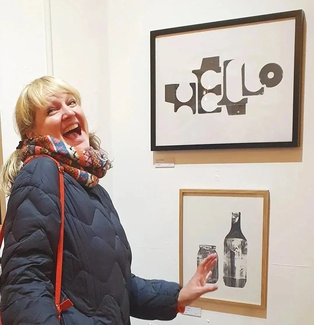 Woman with blonde hair, wearing a black quilted jacket and colorful scarf, standing in an art gallery, smiling and pointing at two framed artworks on the white wall behind her.