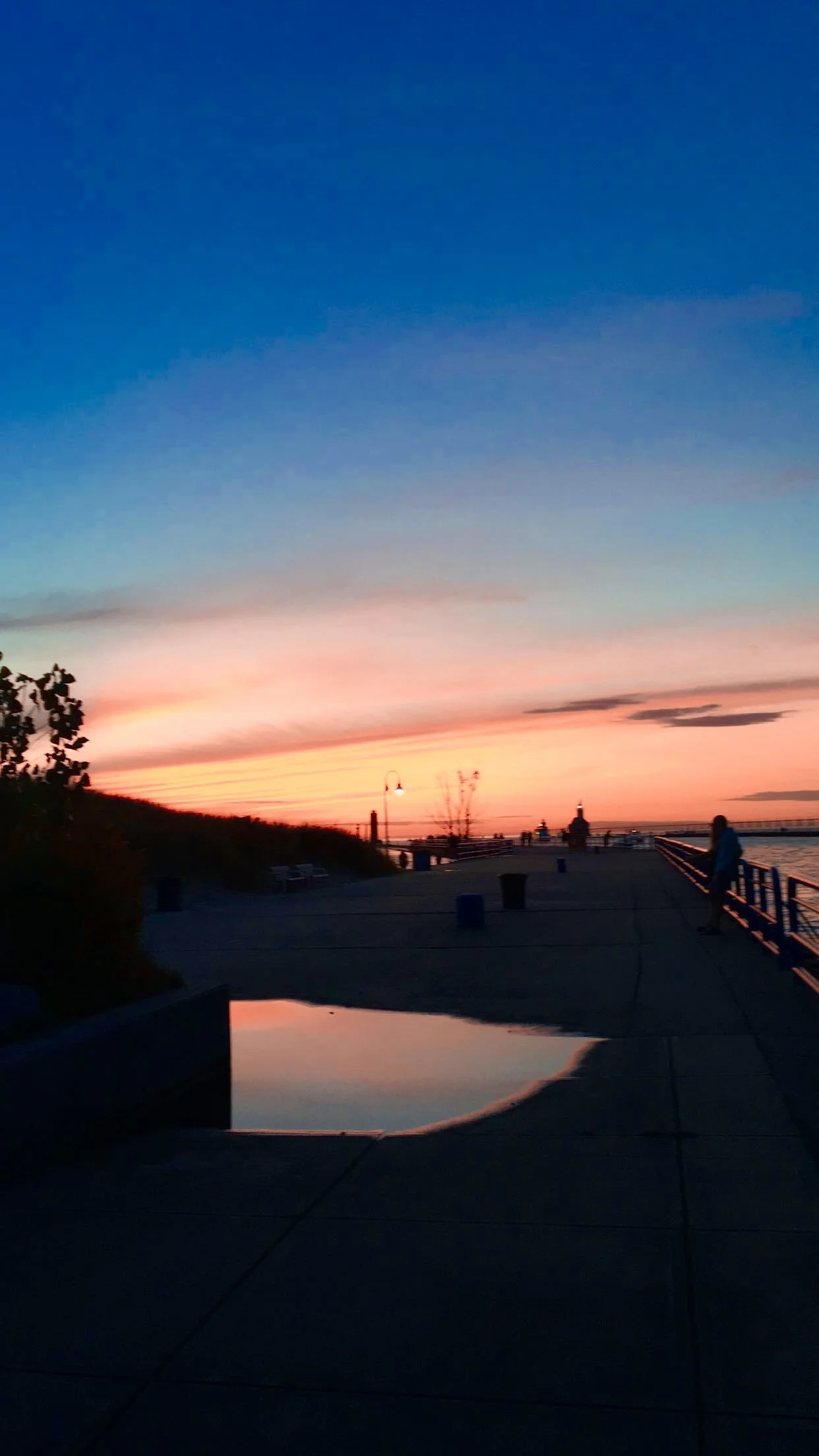 Sunset over a pier with a puddle reflecting the sky, people walking, and street lamps along the path.