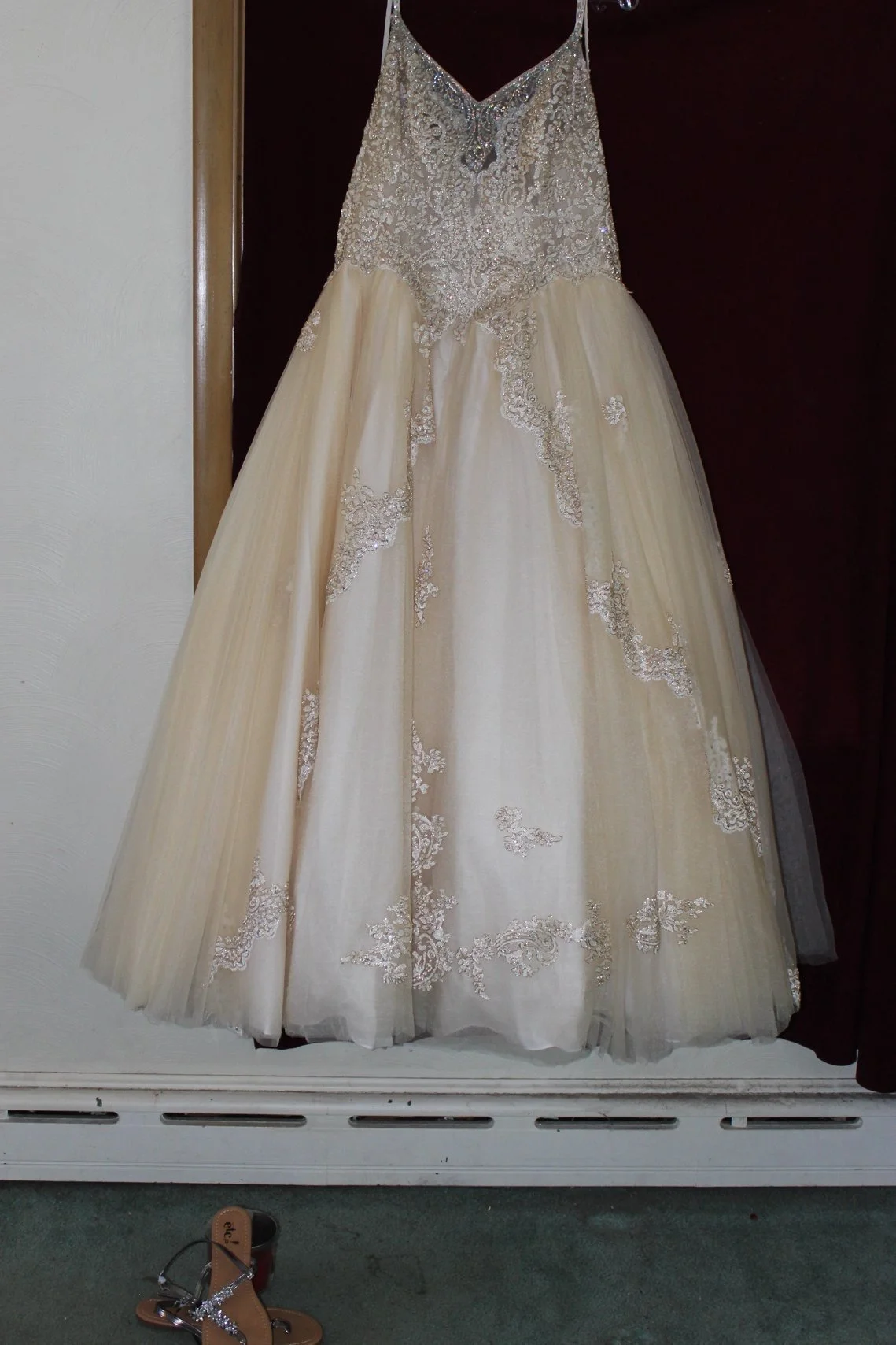 A wedding dress with intricate lace and beadwork, hanging on a dark wooden background, with a pair of brown sandals with an ankle strap and rhinestone embellishments on the floor in front.