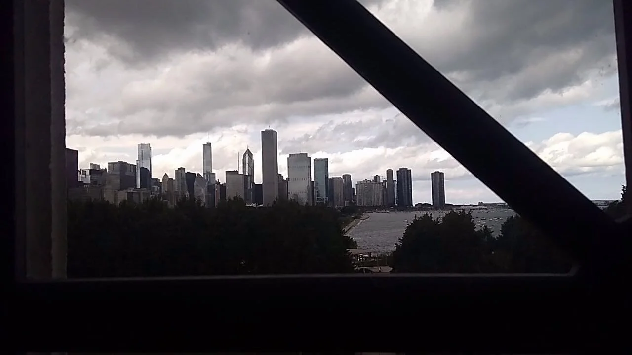 View of a city skyline with tall skyscrapers under a cloudy sky, seen through a window with black framing and diagonal support beams, near a water body and trees.