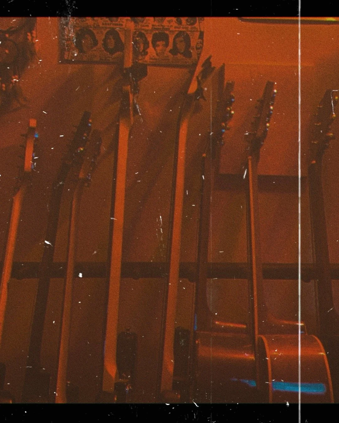A collection of six guitars hanging on a wall and a poster with nine black-and-white headshots above them, with a reddish background and some scattered light reflections.