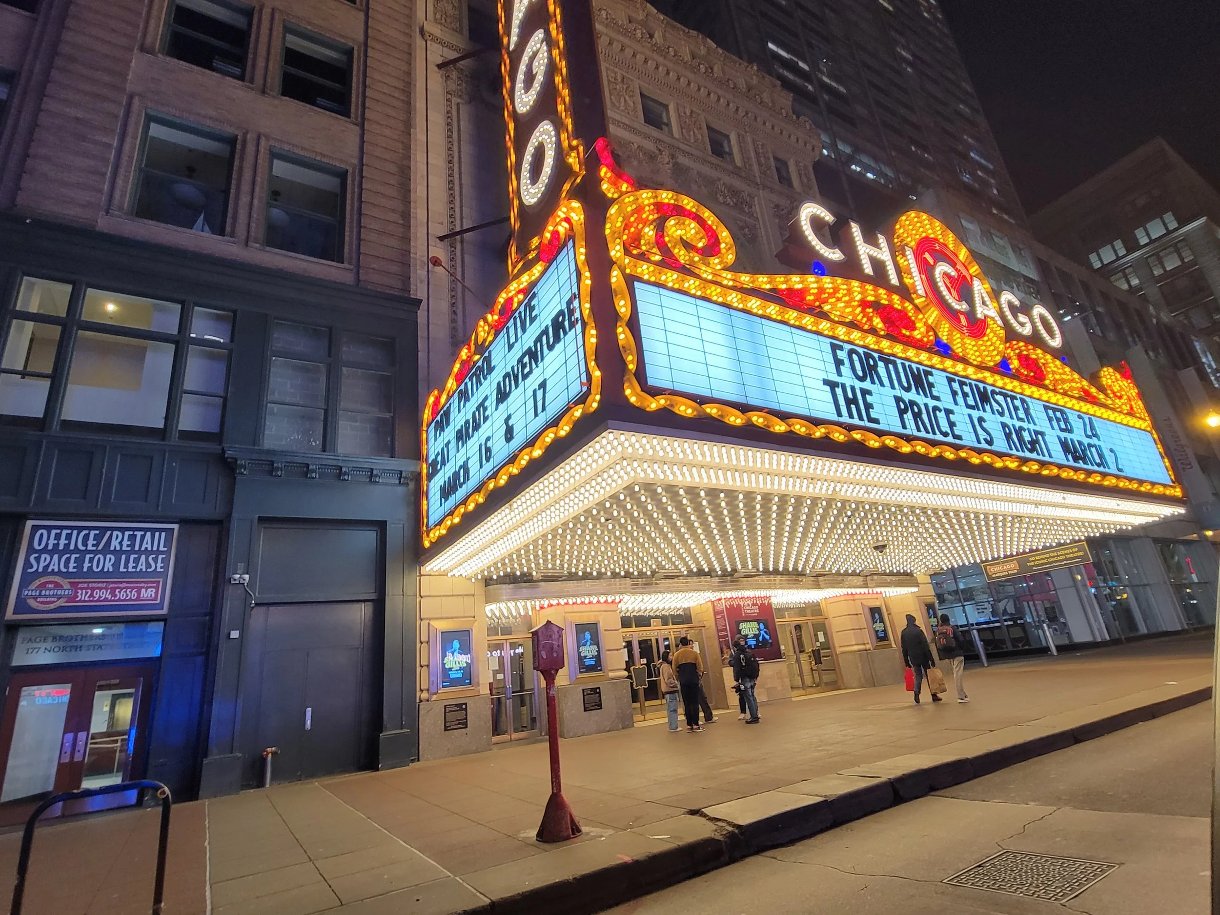 The Chicago theater marquee lit up at night with bright lights, displaying the word 'Chicago' and event information on a marquee board with a few people standing and walking nearby.