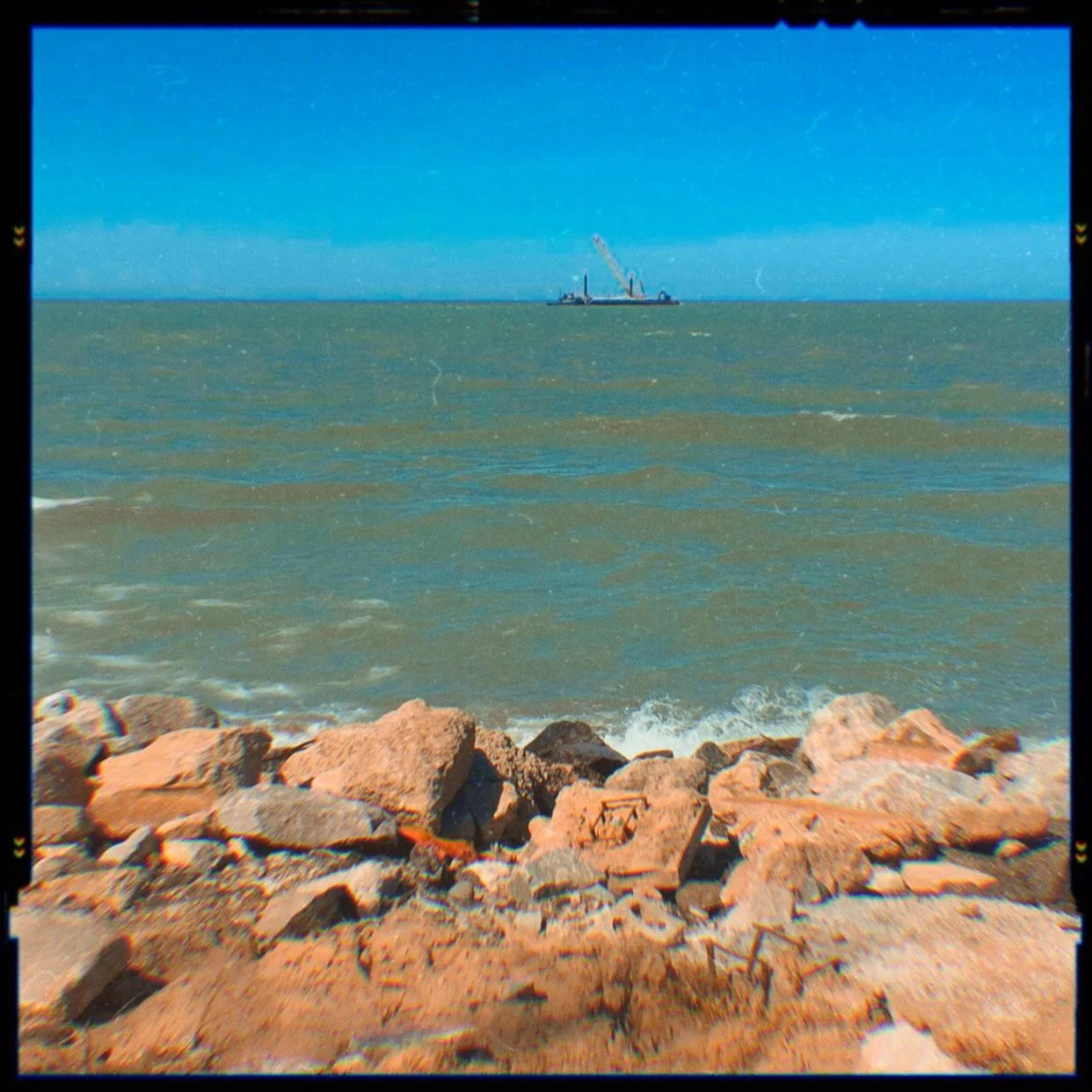 View of the ocean with rocks in the foreground and a construction barge with a crane in the distance under a blue sky.