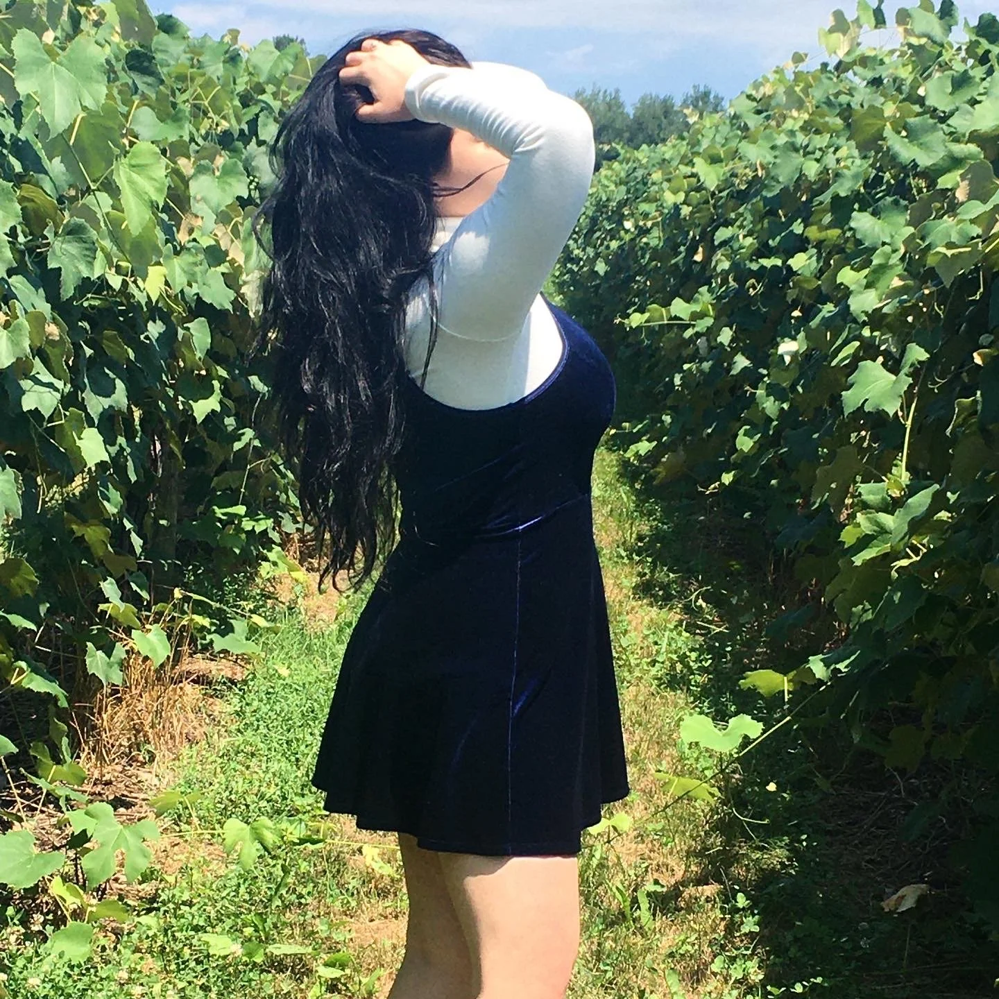 A woman with long black hair, wearing a white long sleeve top under a black velvet dress, standing on a path between green bushy plants outdoors in sunny weather.