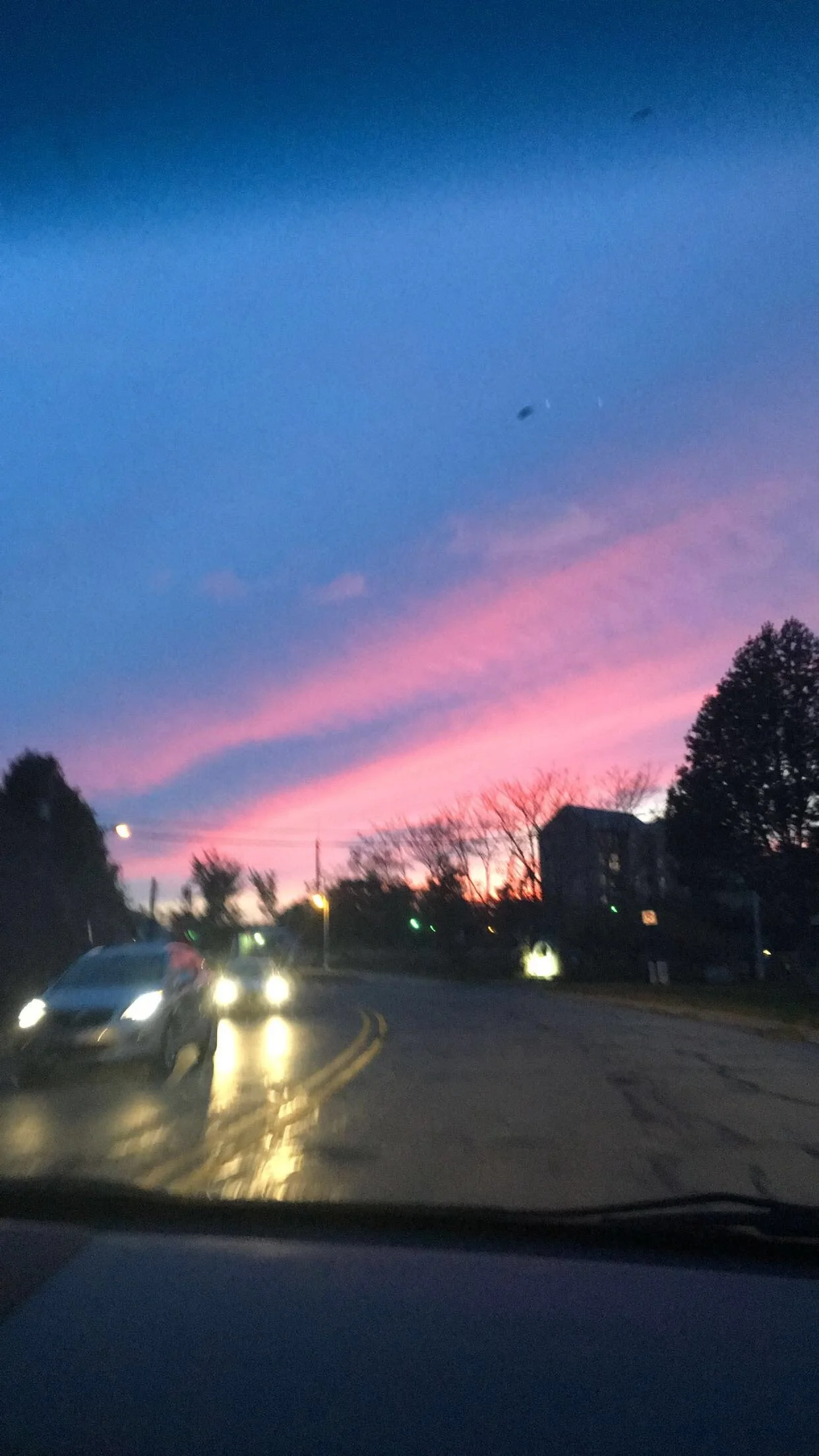 A colorful sunset sky with pink and purple clouds over a street with cars and silhouettes of trees and houses.