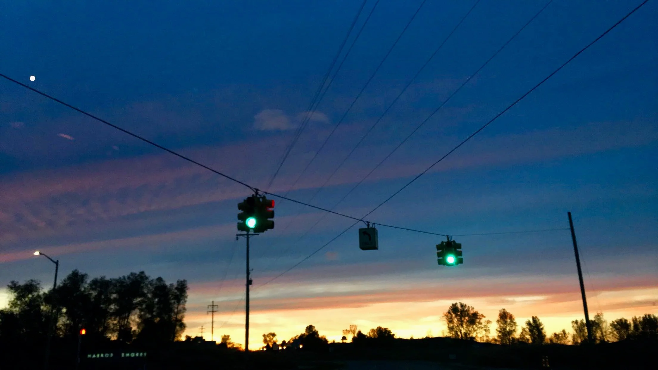 Traffic lights with green signals against a sunset sky with clouds, trees, and power lines.