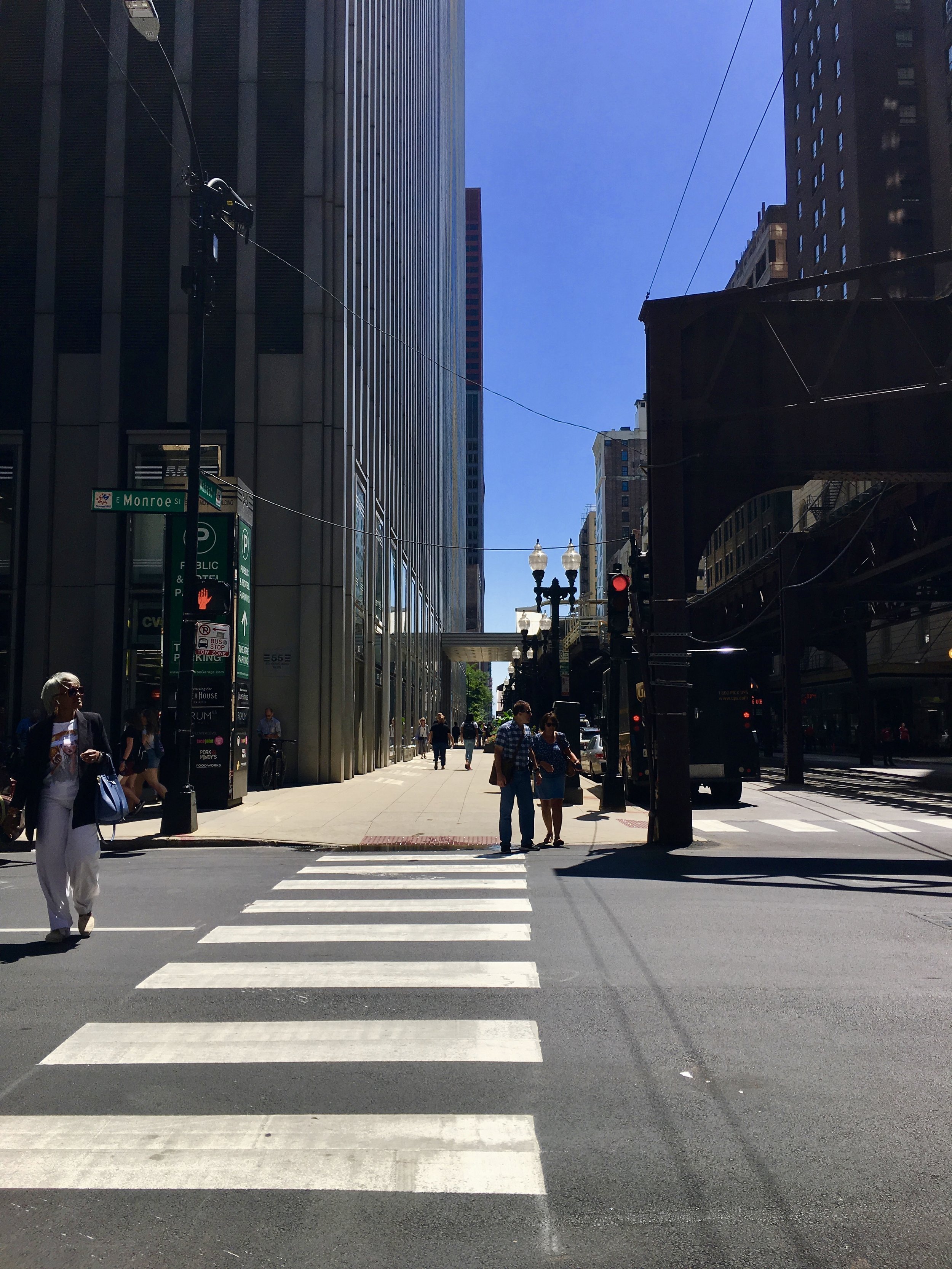 City street scene with crosswalk, pedestrians, tall buildings, traffic signals, and a blue sky.