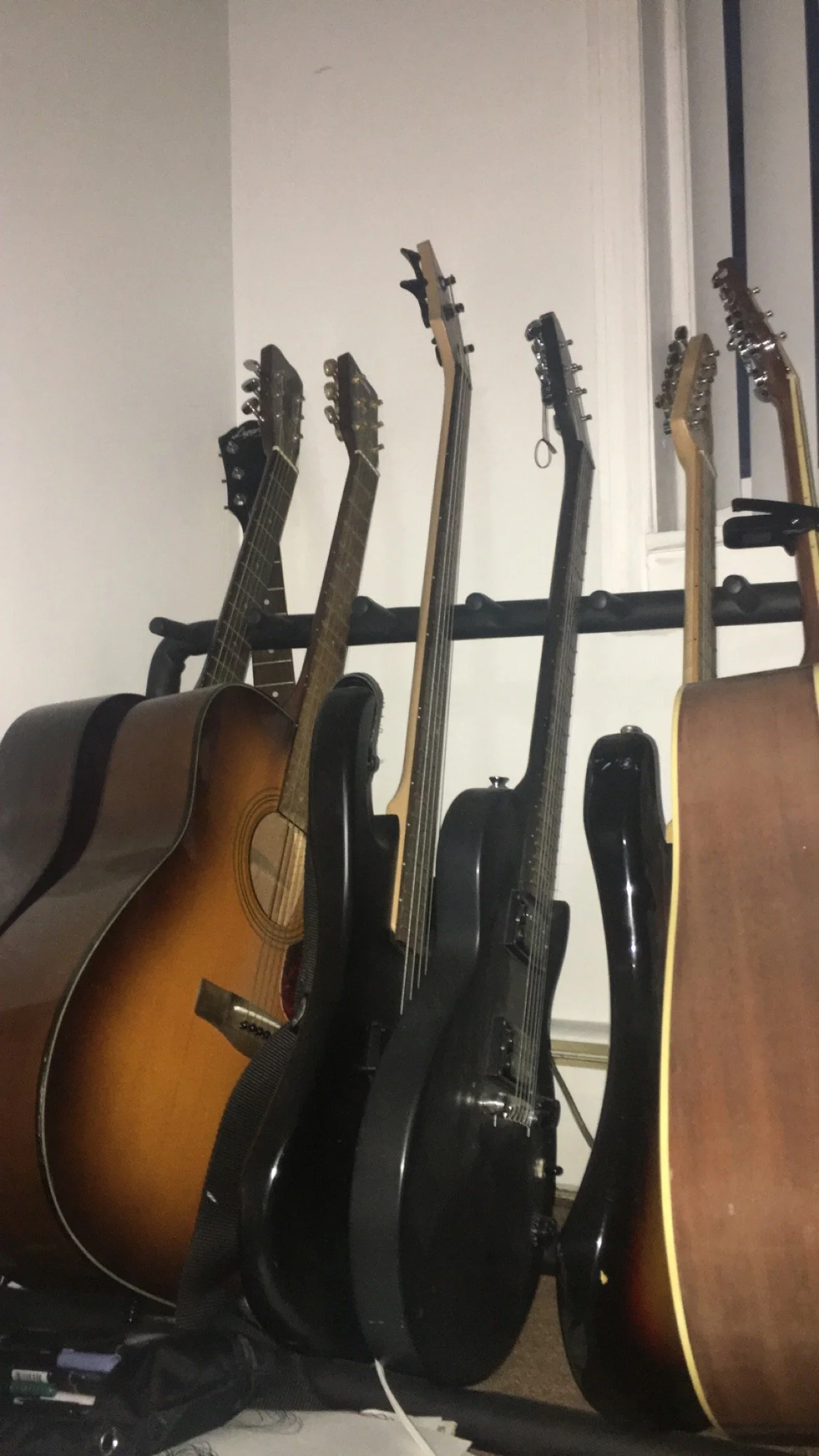 A collection of six guitars standing on a rack in front of a white wall, including acoustic and electric guitars.