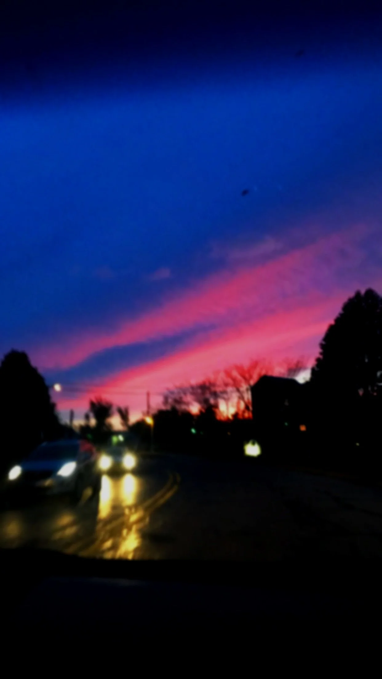Sky at sunset with pink and purple clouds, silhouetted trees and houses, cars with headlights on on a wet road
