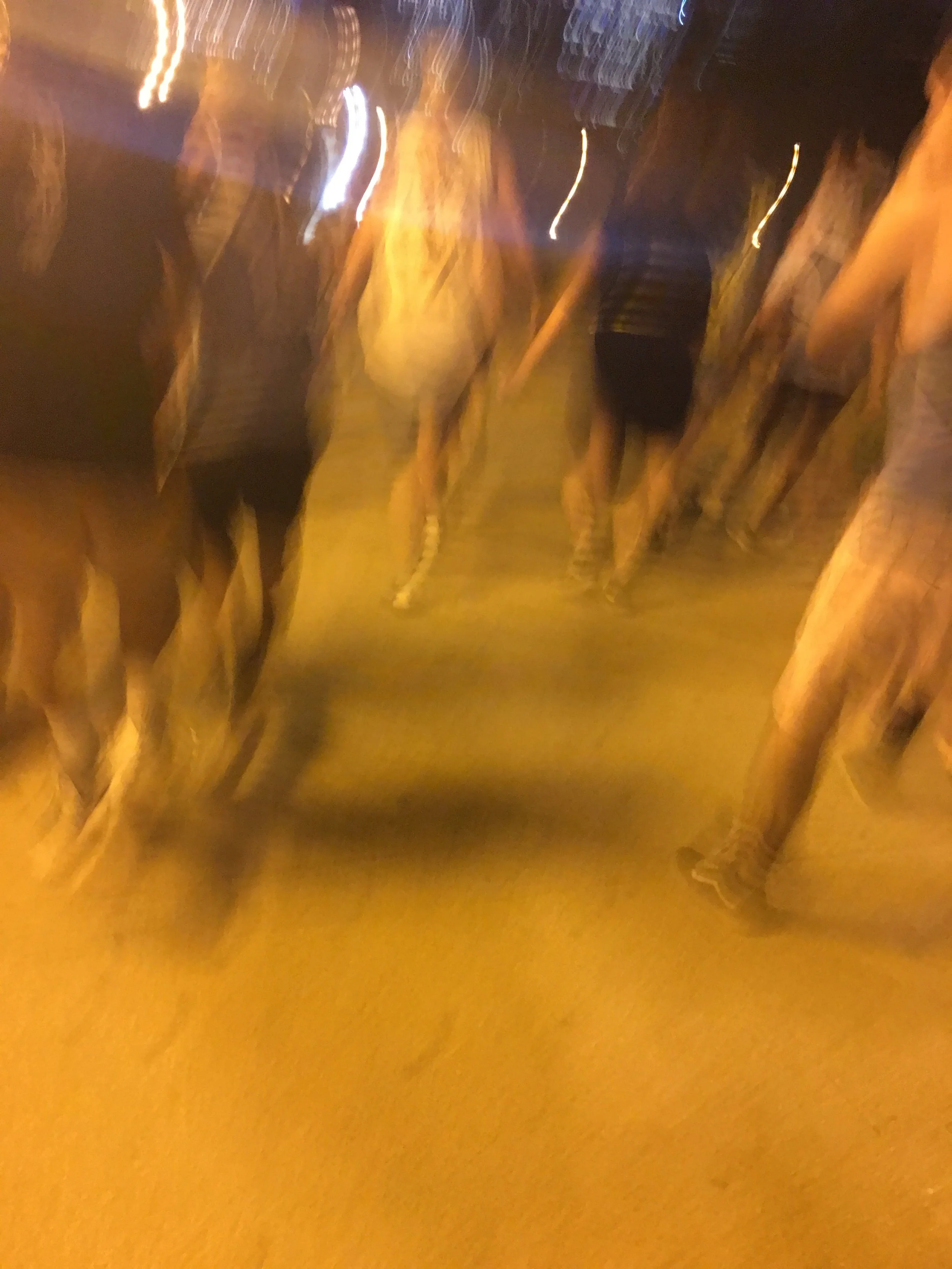 Blurred photo of several people walking at night on a city street with bright lights.