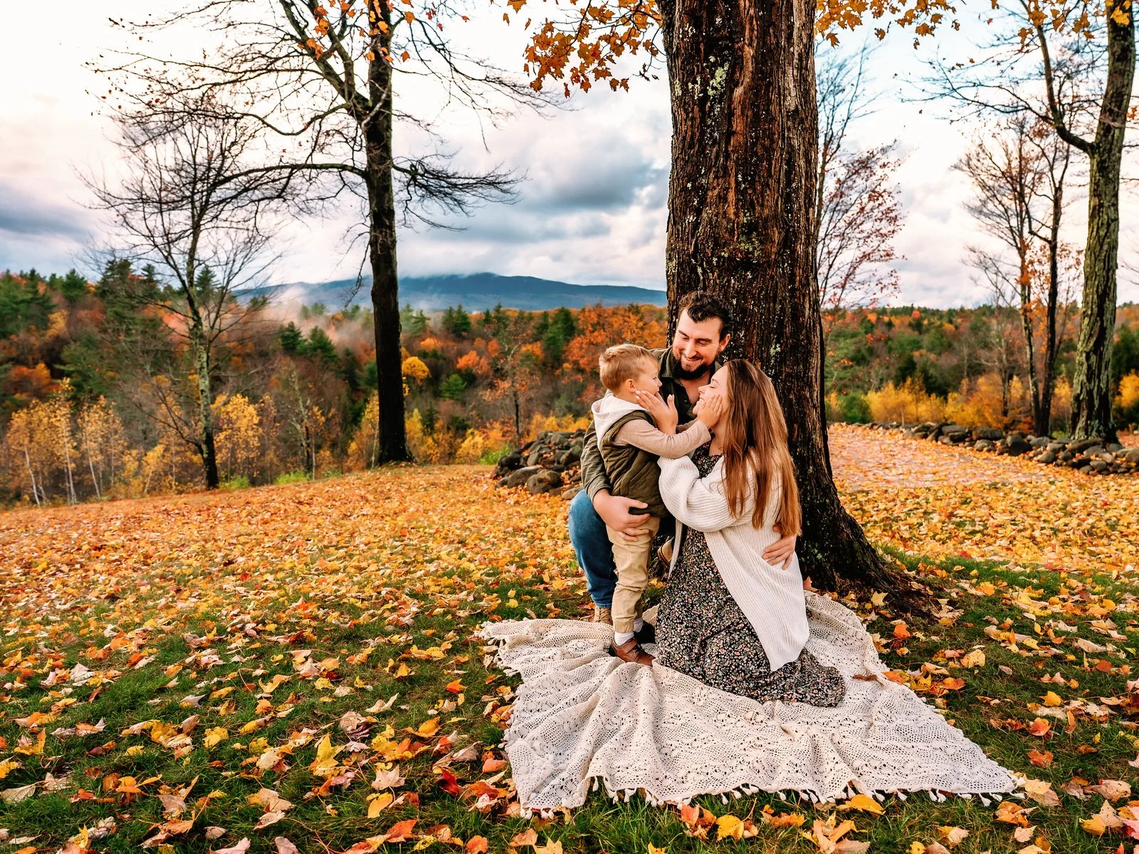 Family of three having a joyful moment outdoors in autumn, with falling leaves, under a large tree, as mountains and colorful trees are visible in the background.