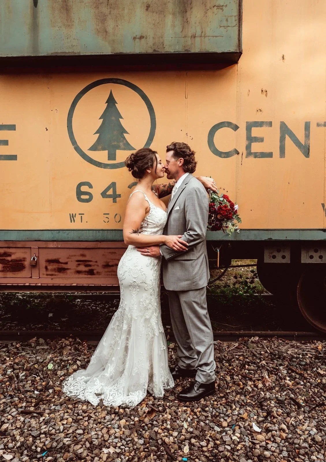 A bride and groom embrace and kiss in front of a vintage orange train car, with the bride in a white lace wedding dress and the groom in a light gray suit holding a bouquet of red, purple, and green flowers.