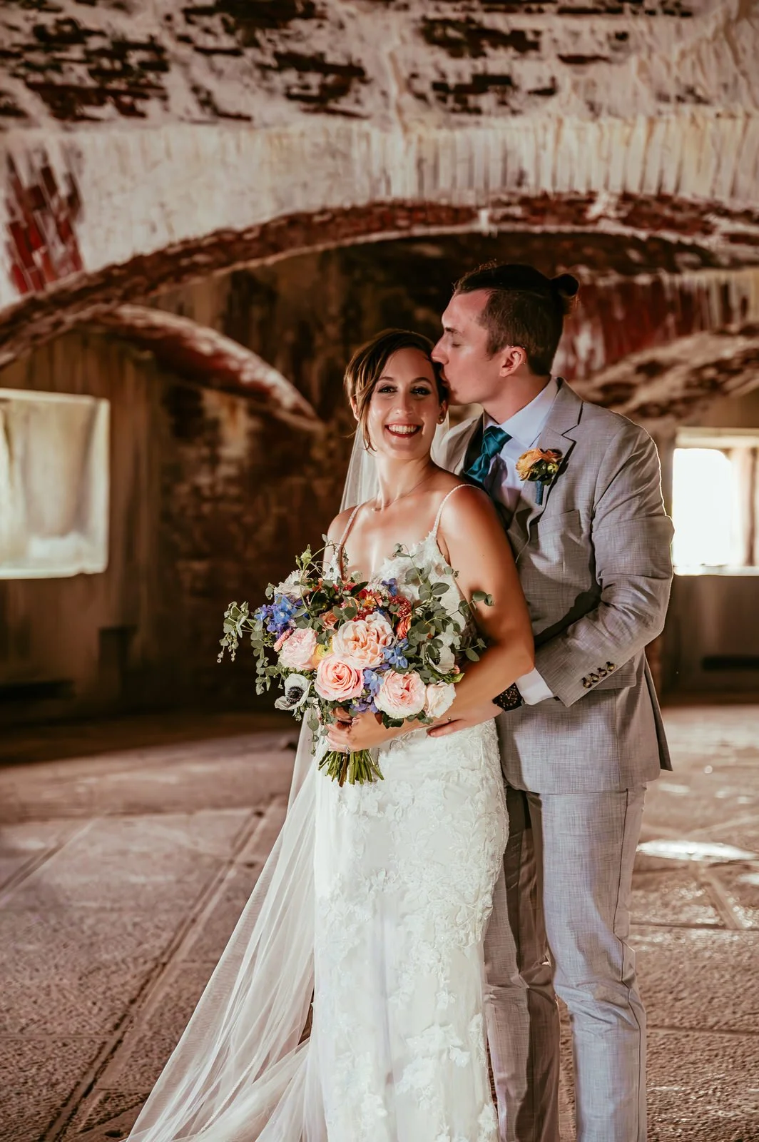 A bride and groom in wedding attire standing inside a rustic brick building, with the groom kissing the bride on her temple, the bride smiling while holding a bouquet of flowers.