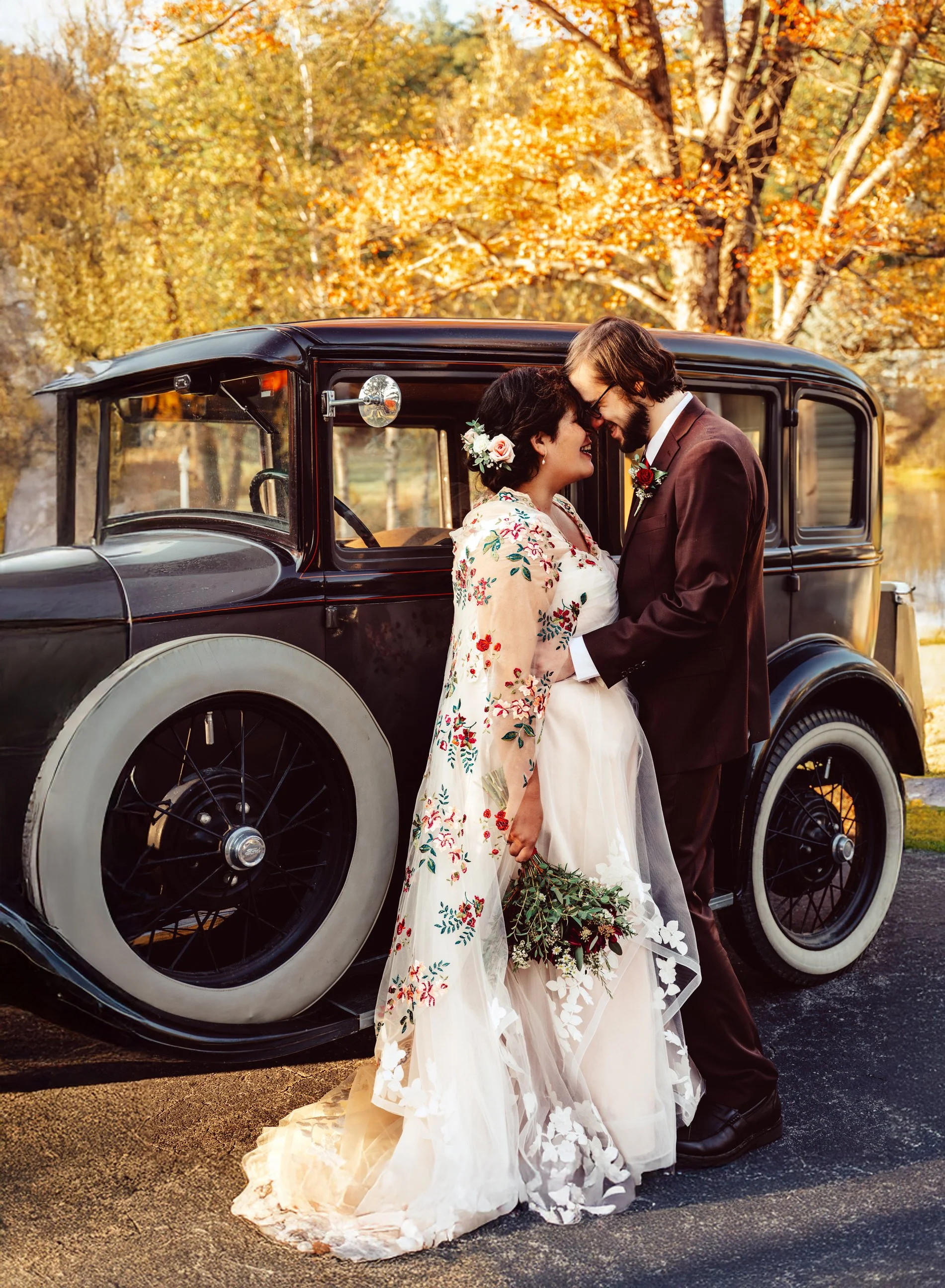 A bride and groom share a tender moment in front of a vintage black car during autumn, surrounded by colorful fall foliage.