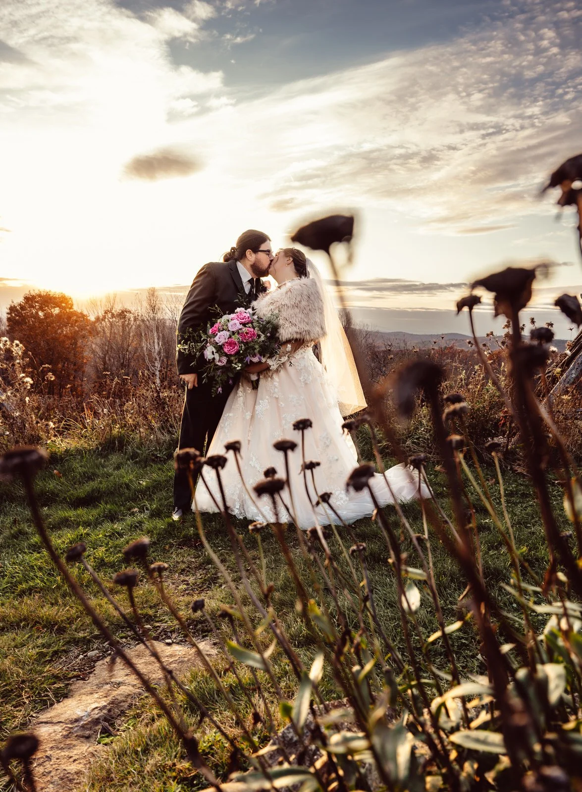 A couple in wedding attire sharing a kiss outdoors during sunset, with wildflowers in the foreground and a scenic landscape in the background.