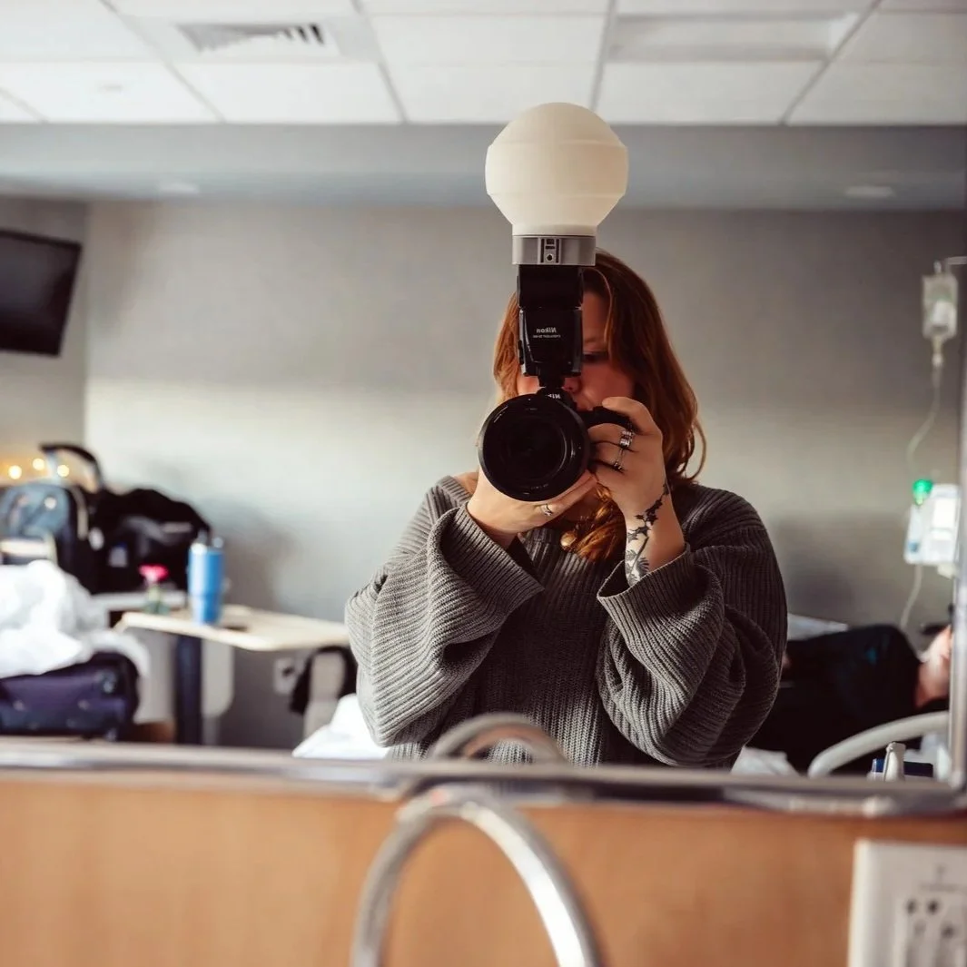 A woman with red hair taking a photo with a camera in a mirror, wearing a gray sweater, in what appears to be a hospital room.