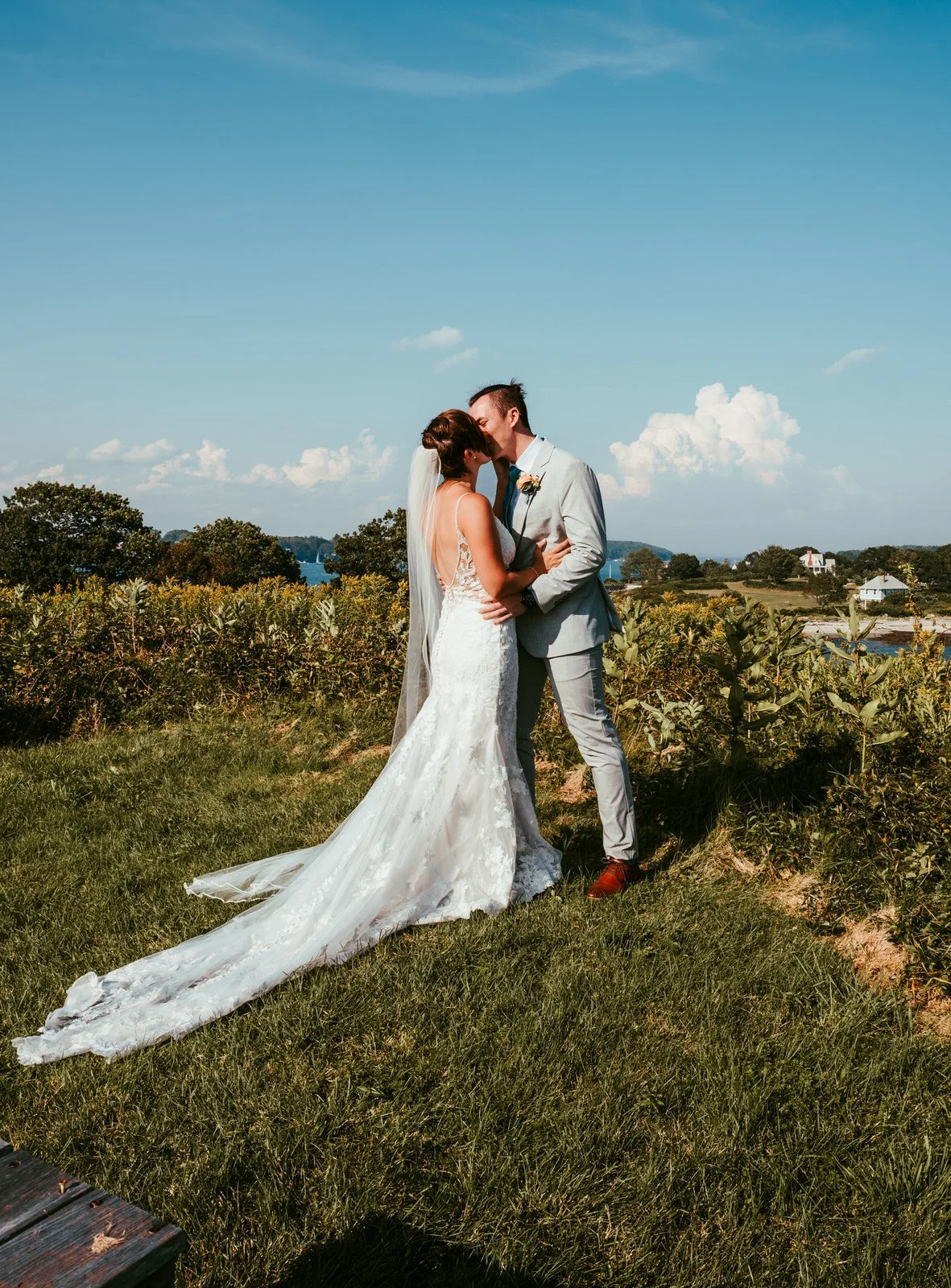 A bride and groom kiss outdoors in a grassy area with trees and some houses in the background on a sunny day.