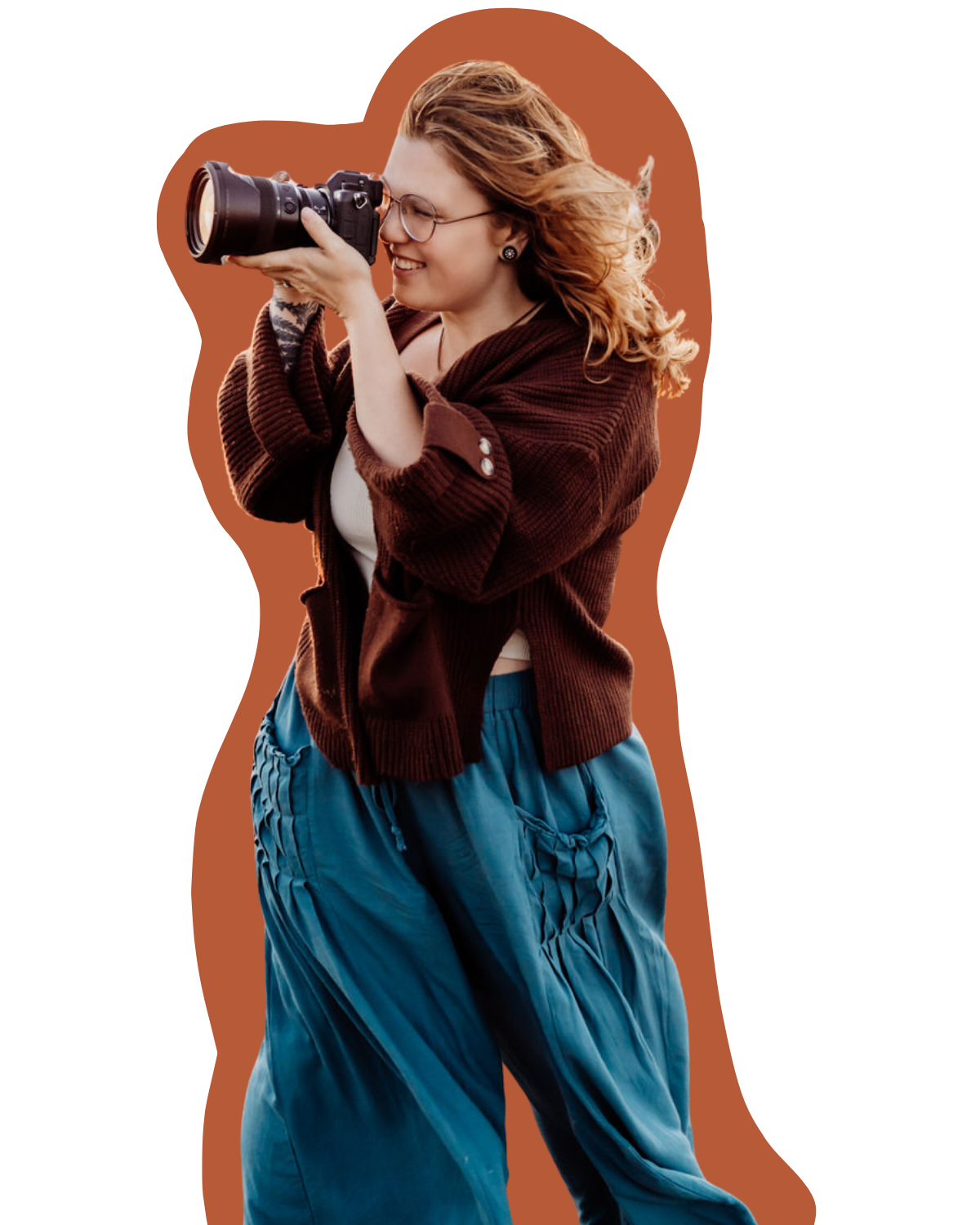 A woman with glasses and curly red hair smiling while looking through a camera's viewfinder, standing outdoors against a brown background.