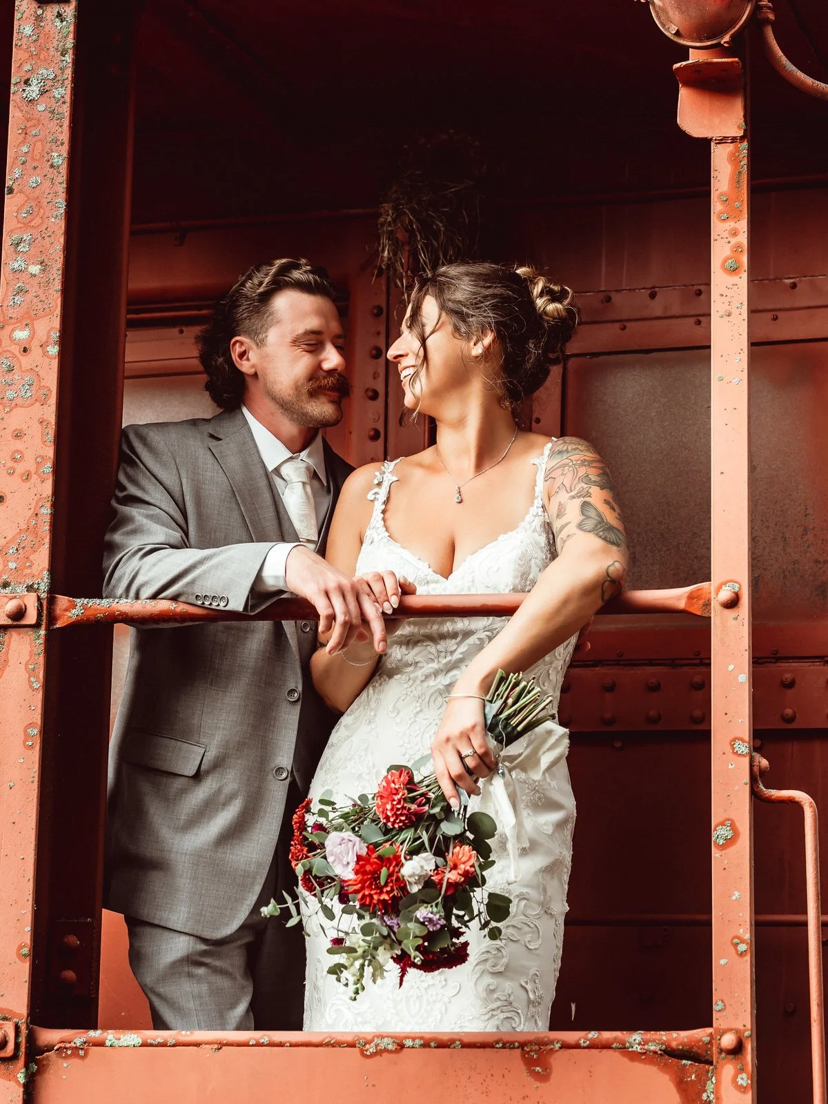 A bride and groom inside a rusty train car, smiling at each other. The bride is holding a bouquet of red and pink flowers, and the groom is wearing a gray suit with a white shirt and tie.