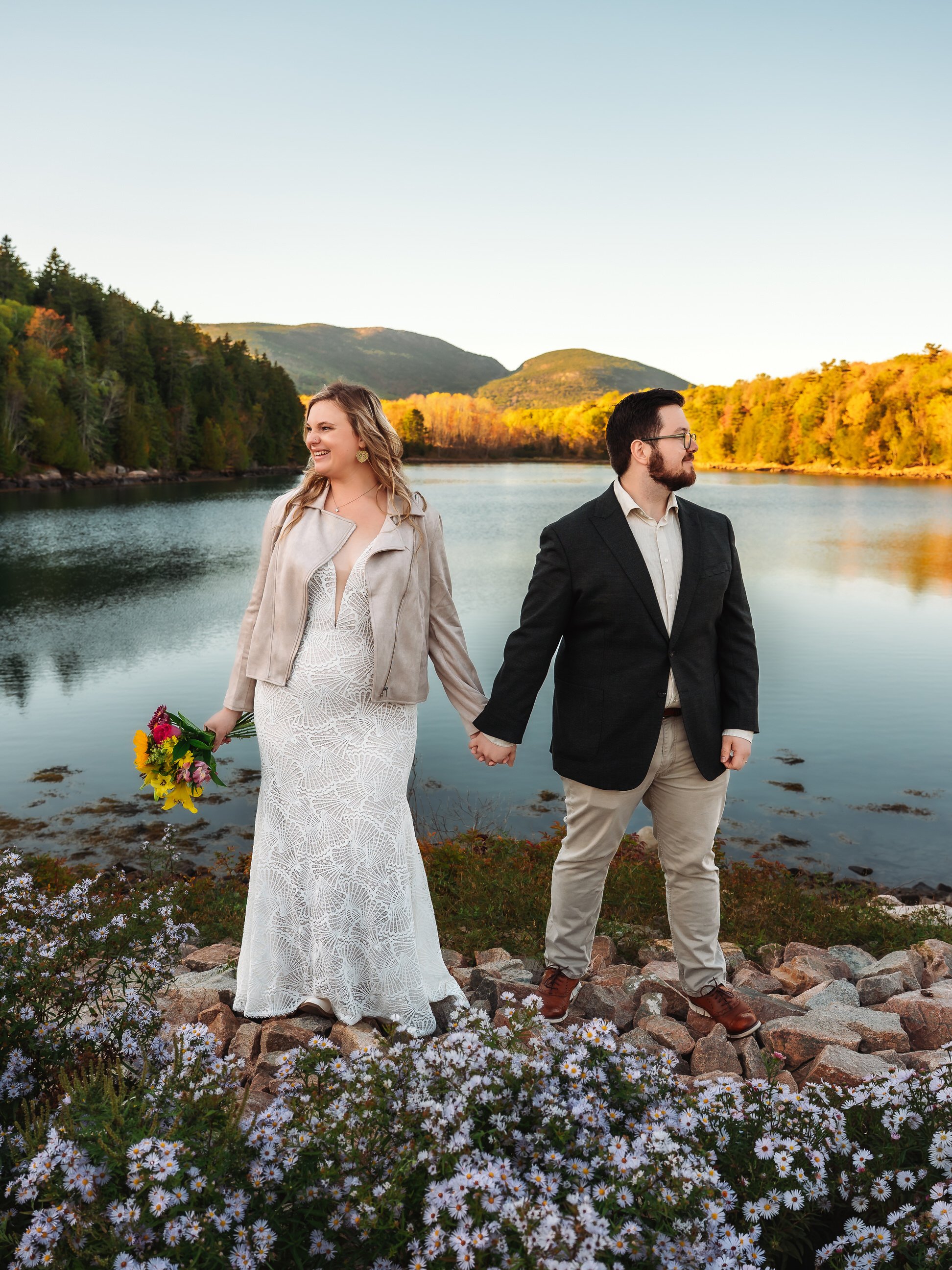 A smiling woman in a white lace dress and beige jacket holding a bouquet, holding hands with a man in a black blazer and beige pants, standing on rocks near a lake with mountains and trees in the background, during sunset.