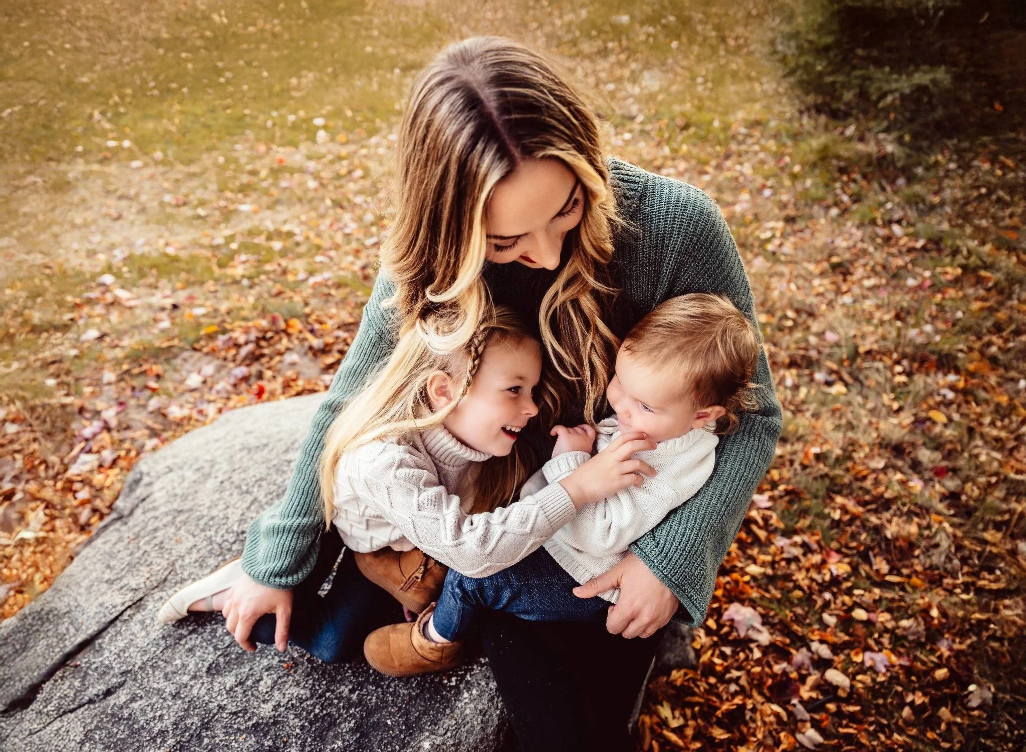A woman sitting on a large rock outdoors during autumn, playing with two young children among fallen leaves.
