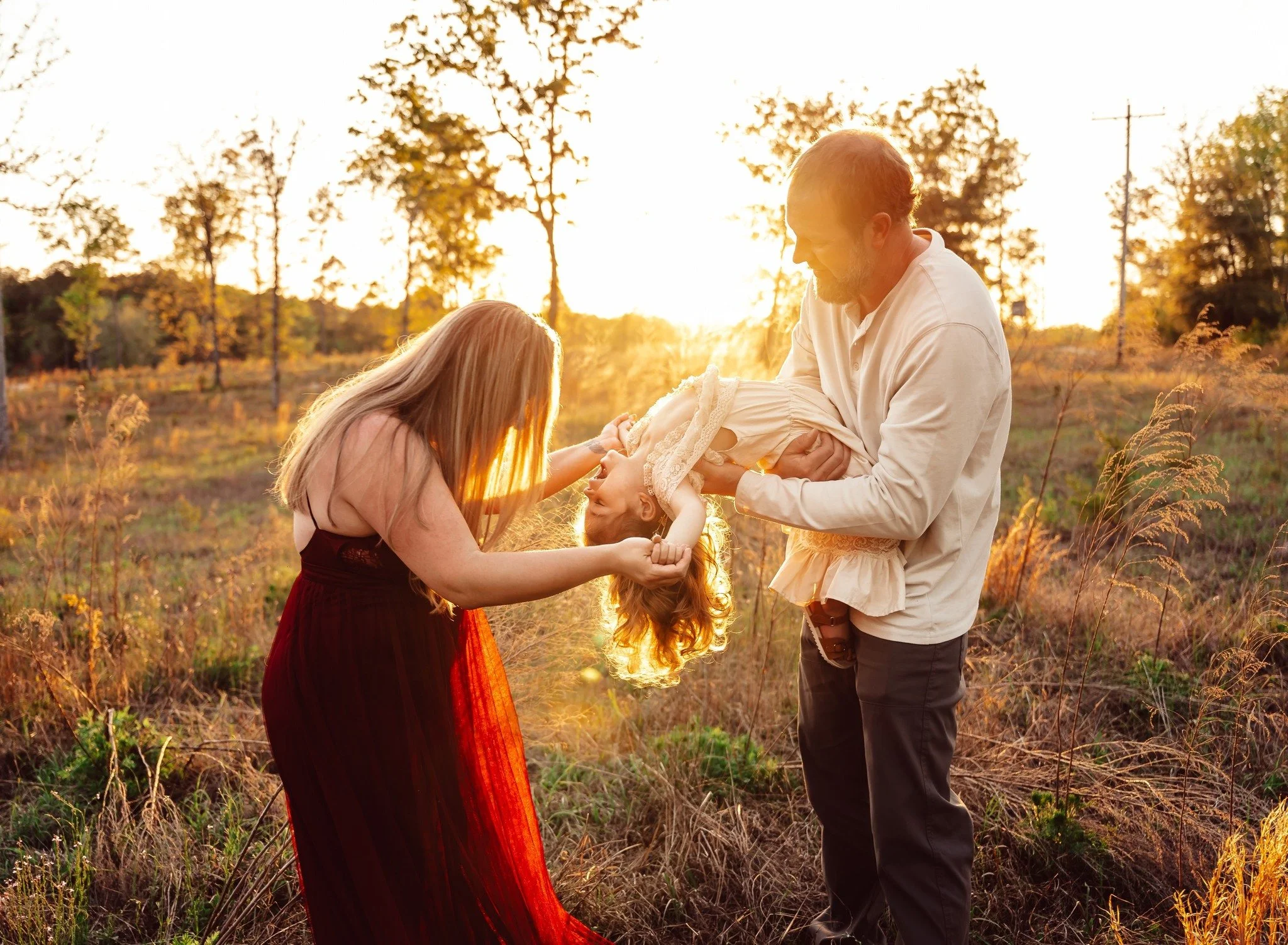 Family of three in a field at sunset, with a man holding a young girl upside down and a woman reaching toward her, all smiling.