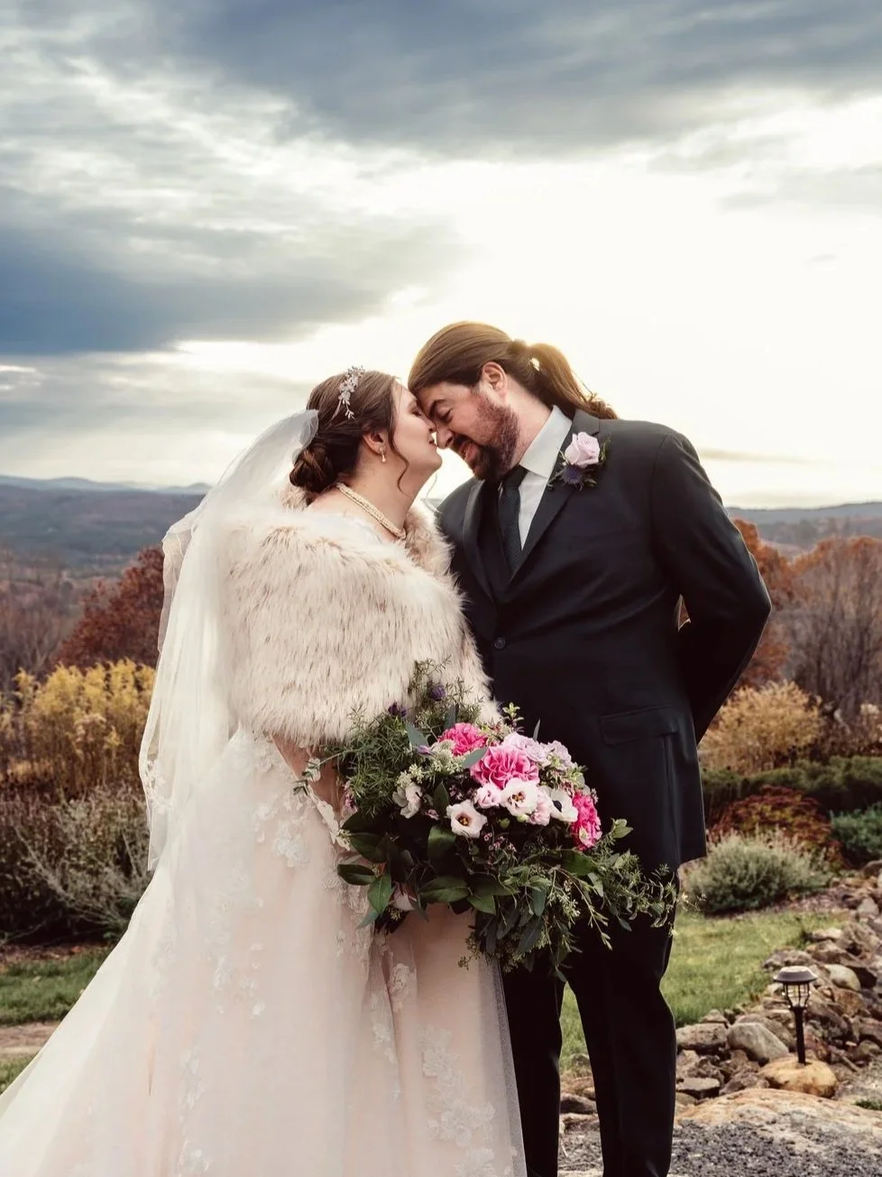A bride and groom sharing a kiss outdoors during their wedding, with a scenic landscape and cloudy sky in the background. The bride is holding a bouquet of pink and purple flowers.
