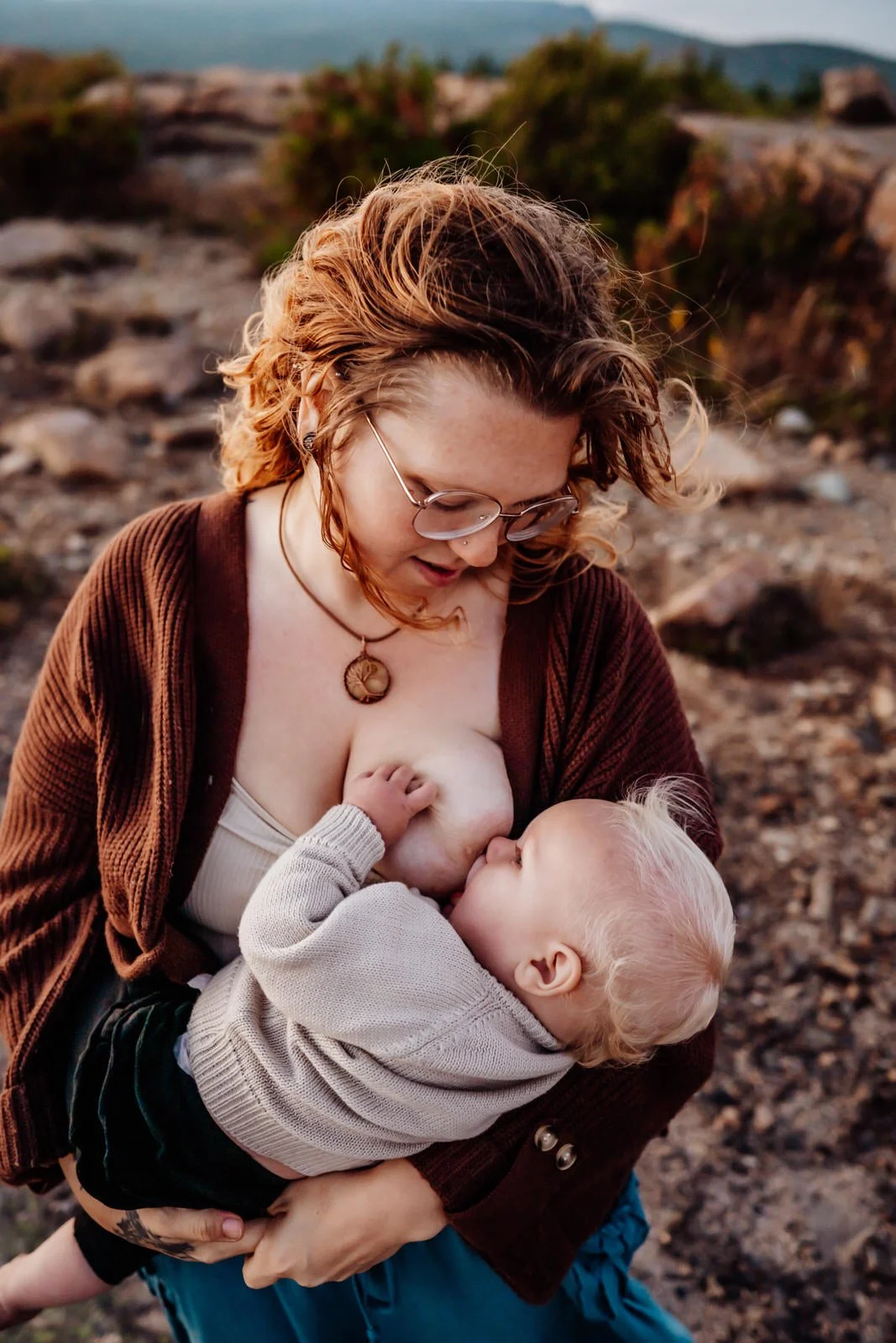 A woman with glasses and curly red hair breastfeeding a young child outdoors in a rocky, mountainous area.