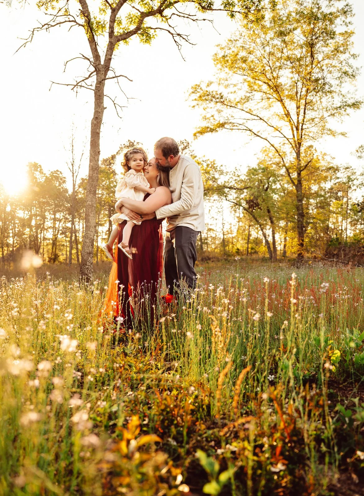 A family of three standing in a grassy field during sunset, with trees in the background. The mother and father are holding their young daughter between them, and they are all smiling and embracing.