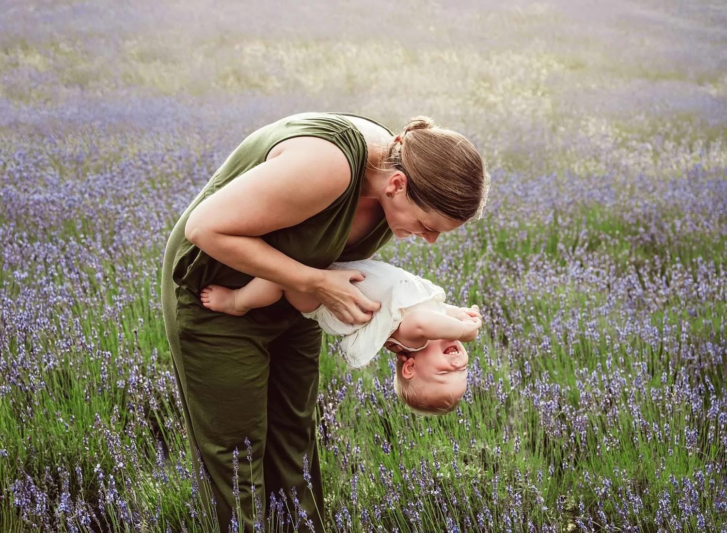 A woman with long brown hair holding a laughing young girl upside down in a lavender field.