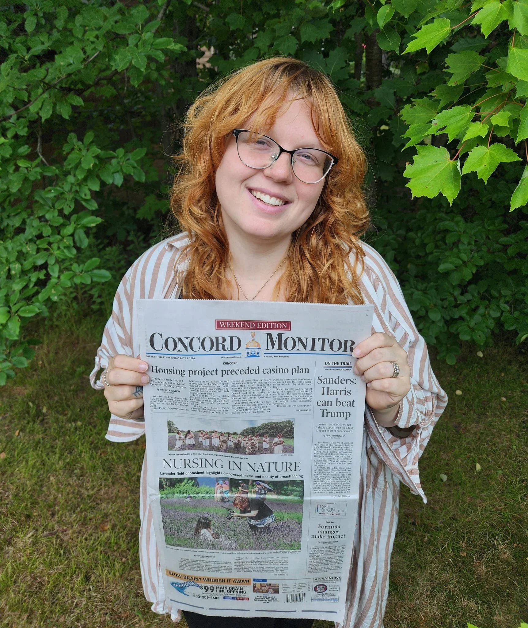 A young woman with red curly hair and glasses holding a newspaper titled "Concord Monitor," standing outdoors in front of green leafy bushes, smiling at the camera.