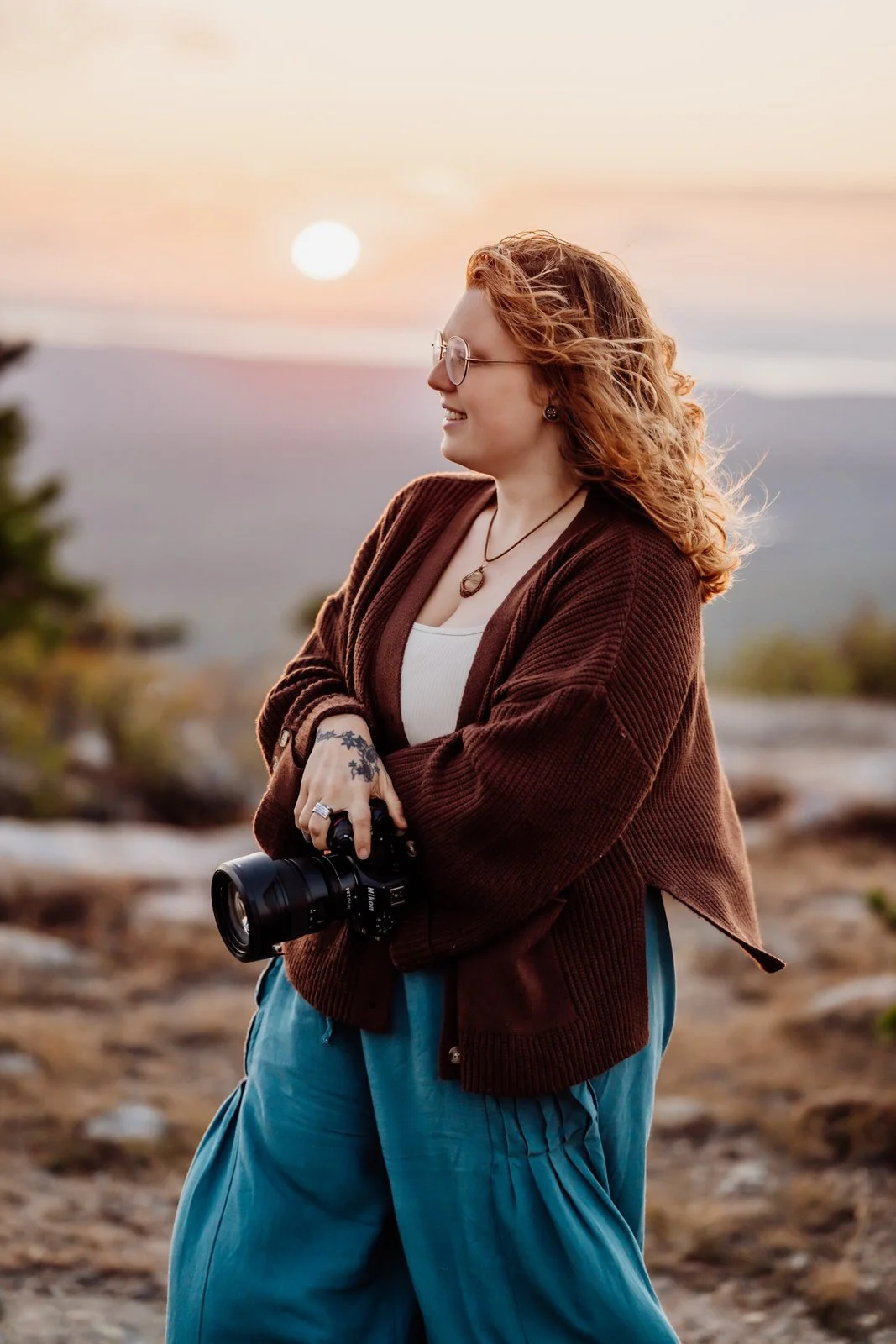 A woman with curly red hair and glasses stands outdoors at sunset, holding a camera and smiling.