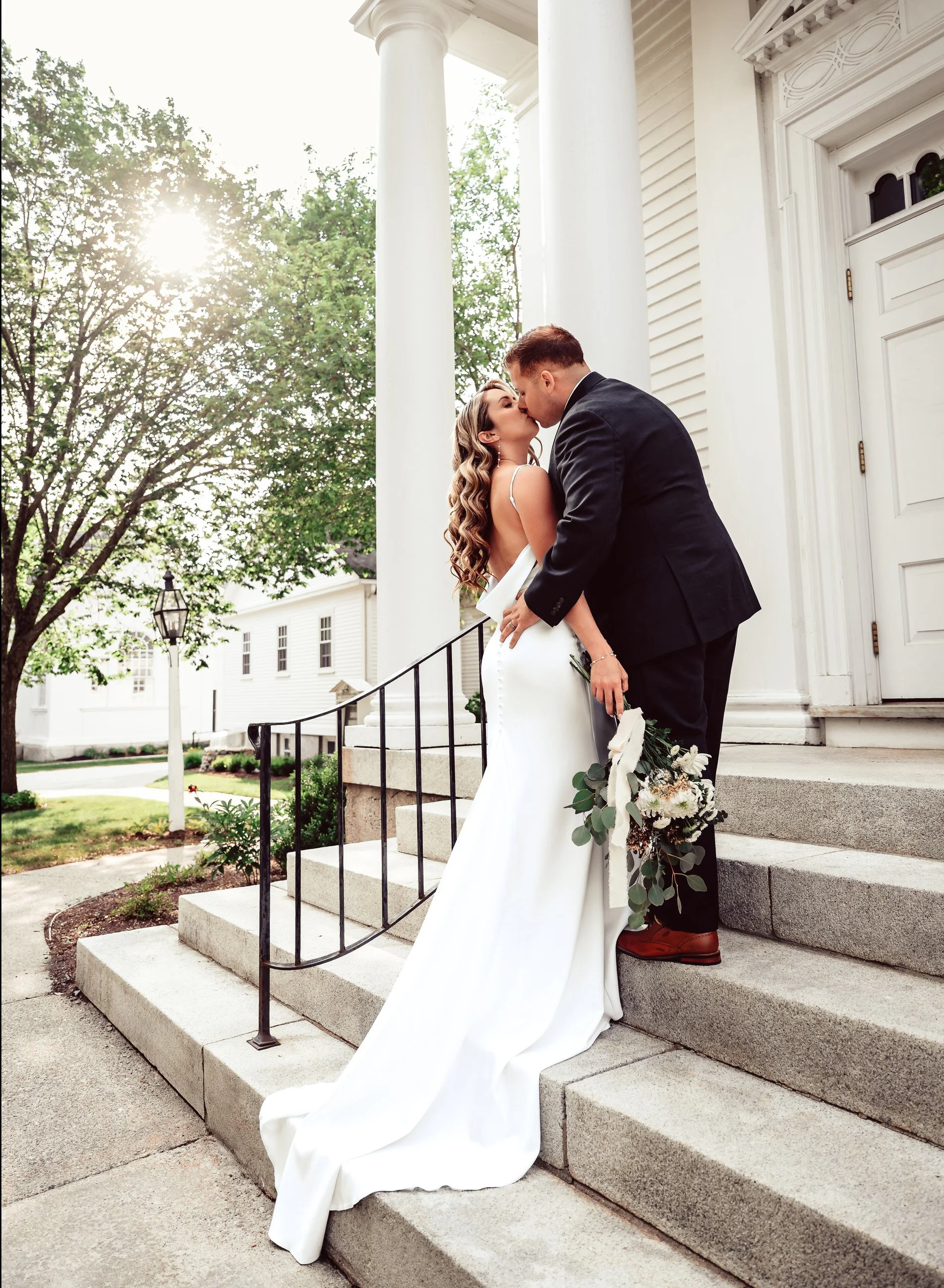 A bride and groom share a kiss on the steps of a white building with large columns and a classical architectural style, with greenery and trees in the background.