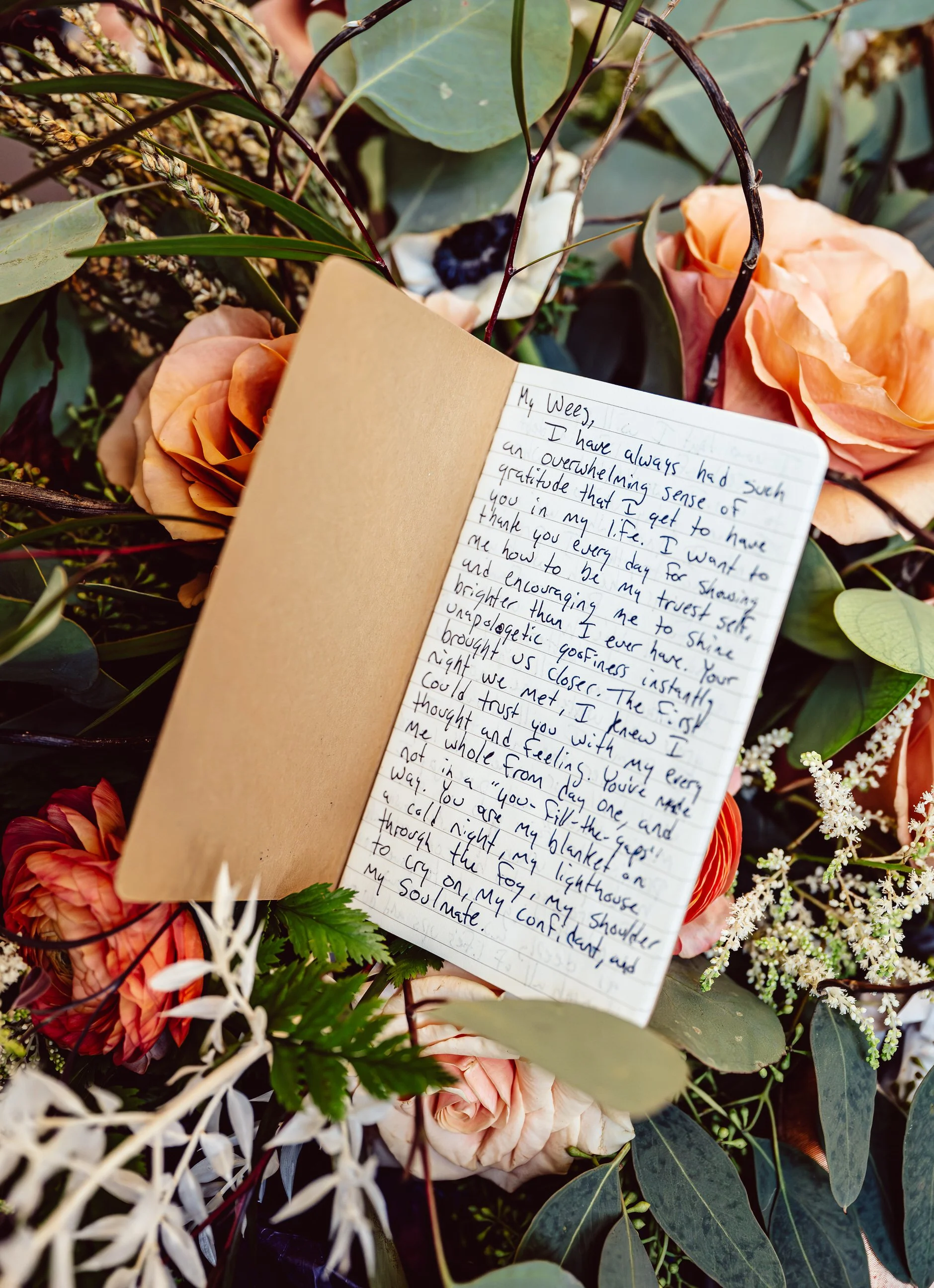 A handwritten note on a small notebook placed among peach-colored roses, greenery, and other flowers in a floral arrangement.