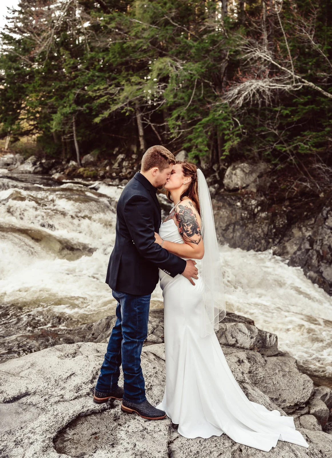 A newlywed couple sharing a kiss on rocks by a rushing river with trees in the background.