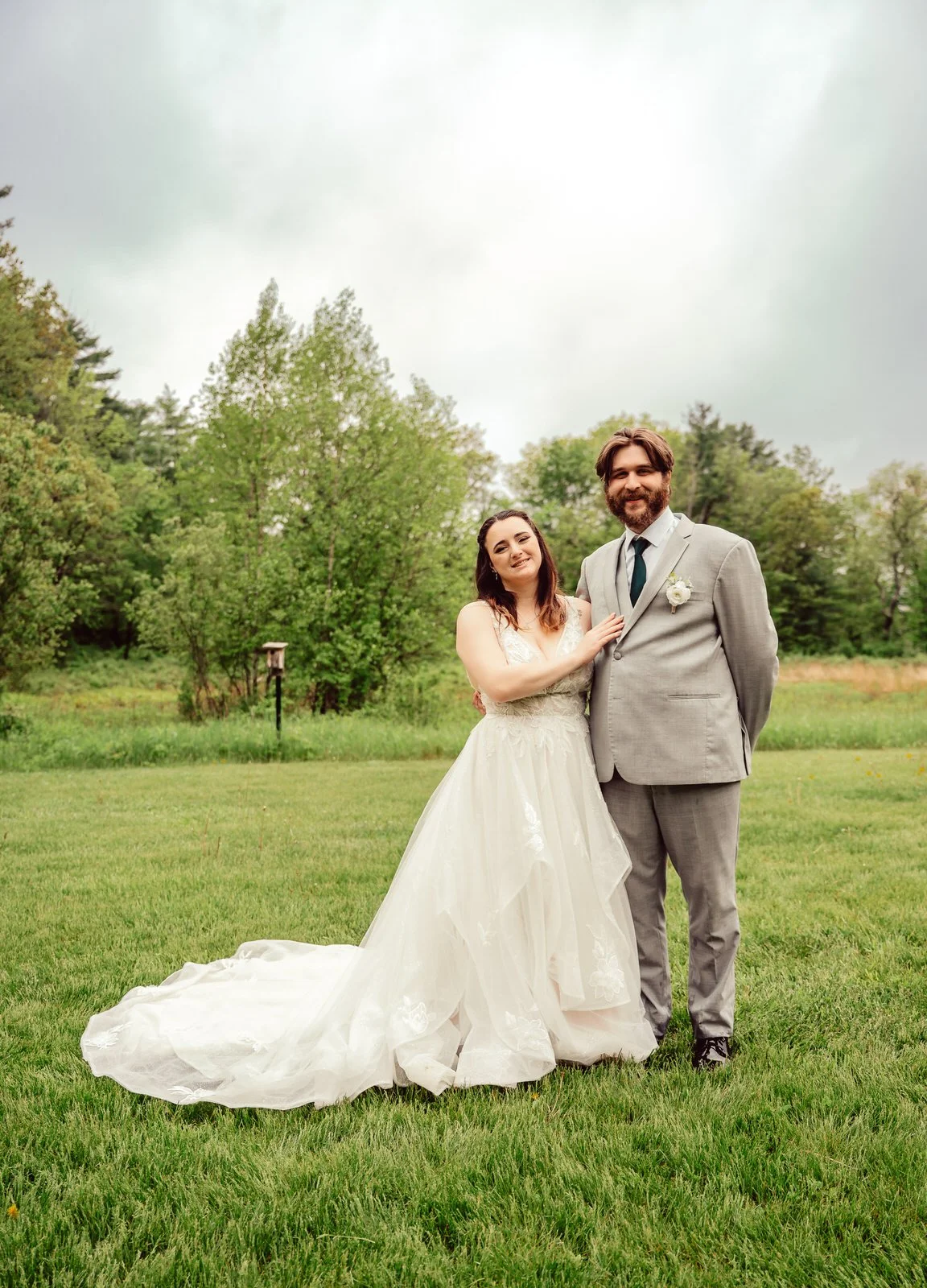 Bride and groom standing together outdoors on a cloudy day, with a green grassy field and trees in the background. The bride is wearing a white wedding gown with a long train, and the groom is dressed in a light gray suit with a white boutonnière.