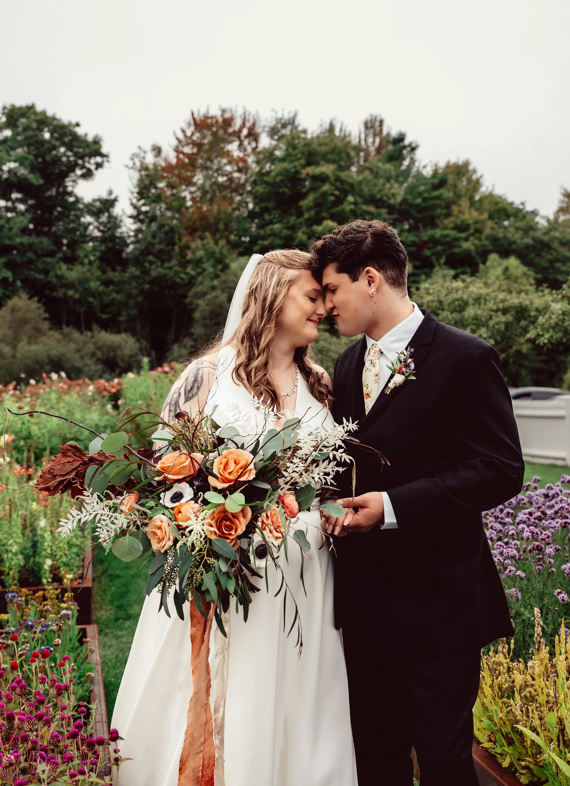 Bride and groom sharing a forehead kiss outdoors in a garden with a variety of colorful flowers and green trees.