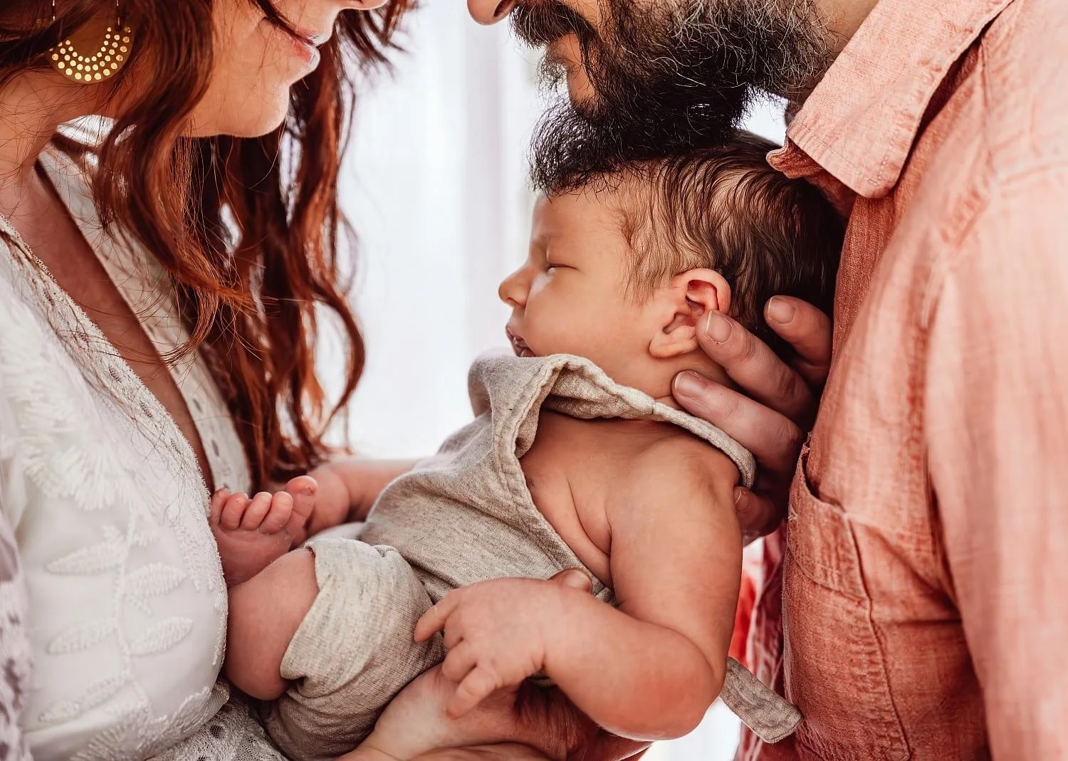 A baby boy being held by his parents, who are looking at him lovingly. The mother has red hair and is wearing earrings, while the father has a beard and is wearing a light-colored shirt. The baby is dressed in a light beige outfit and is sleeping peacefully.