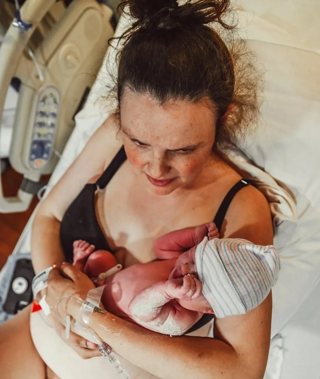 A woman in a hospital bed cradles her newborn baby, who is wearing a striped hat, just after birth.