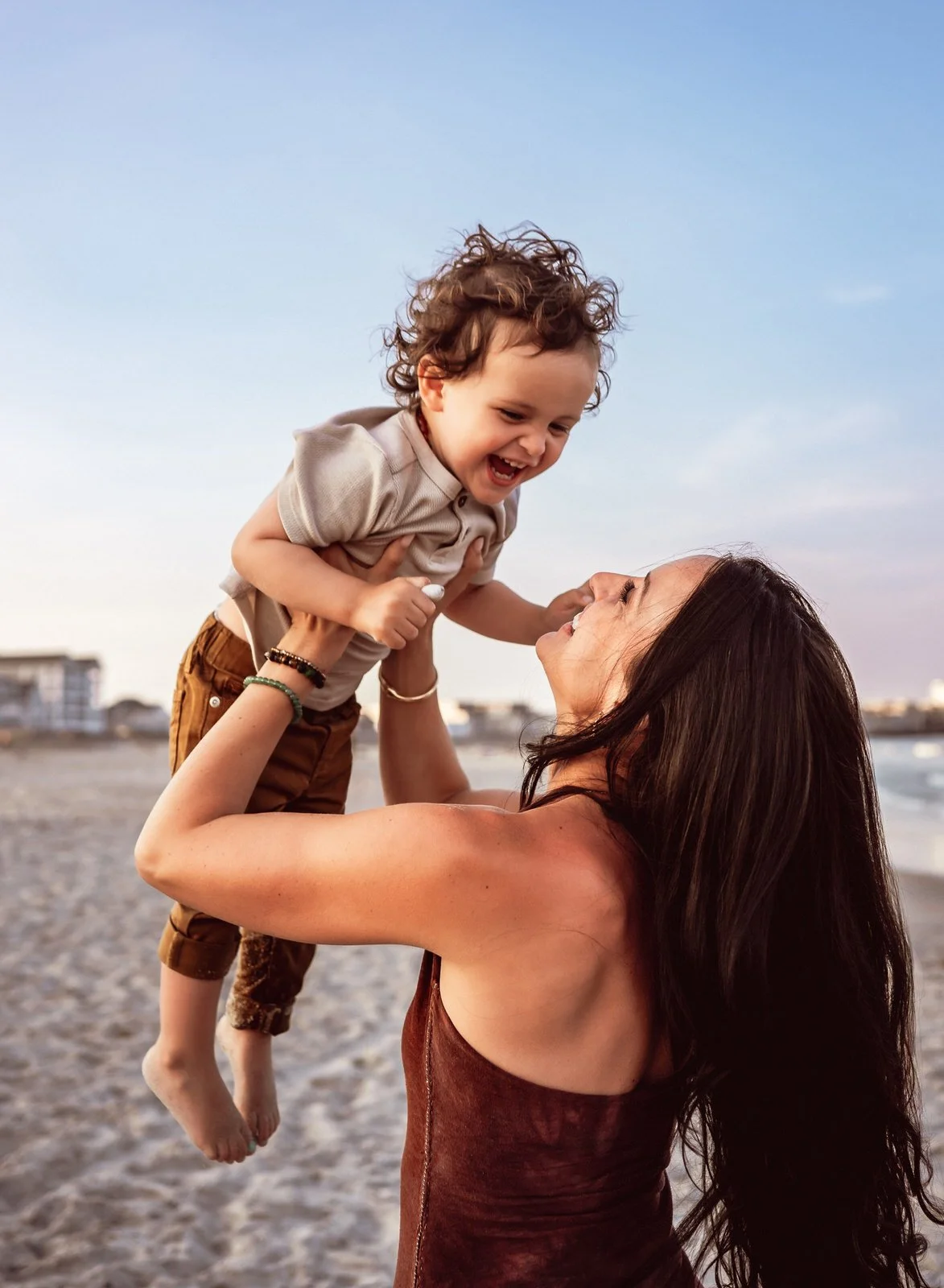 A woman lifting a young boy on a beach with buildings and a cloudy sky in the background.