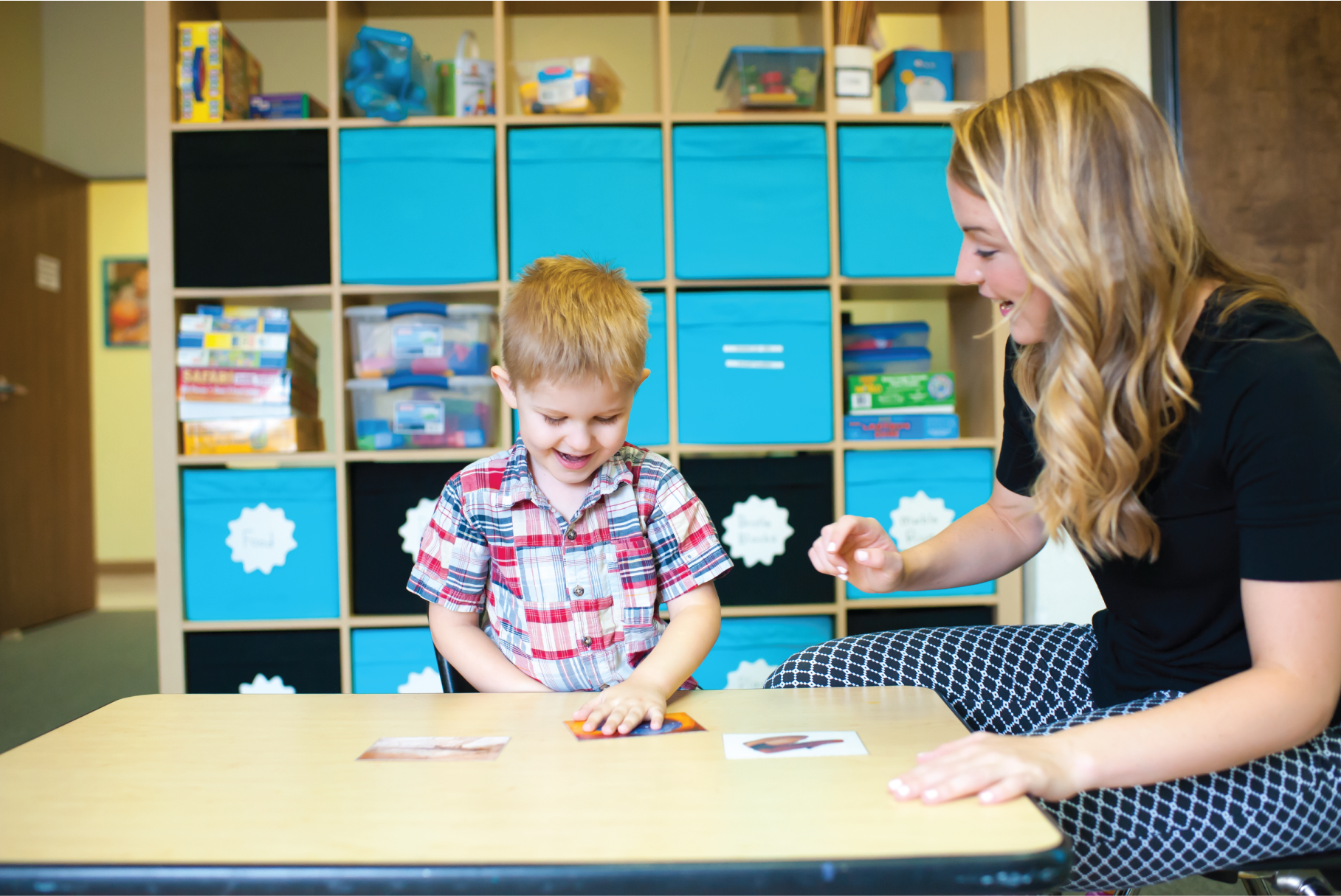 A young boy and a woman sitting at a table in a classroom, playing a game with picture cards, with a cubed storage unit filled with toys and supplies behind them.