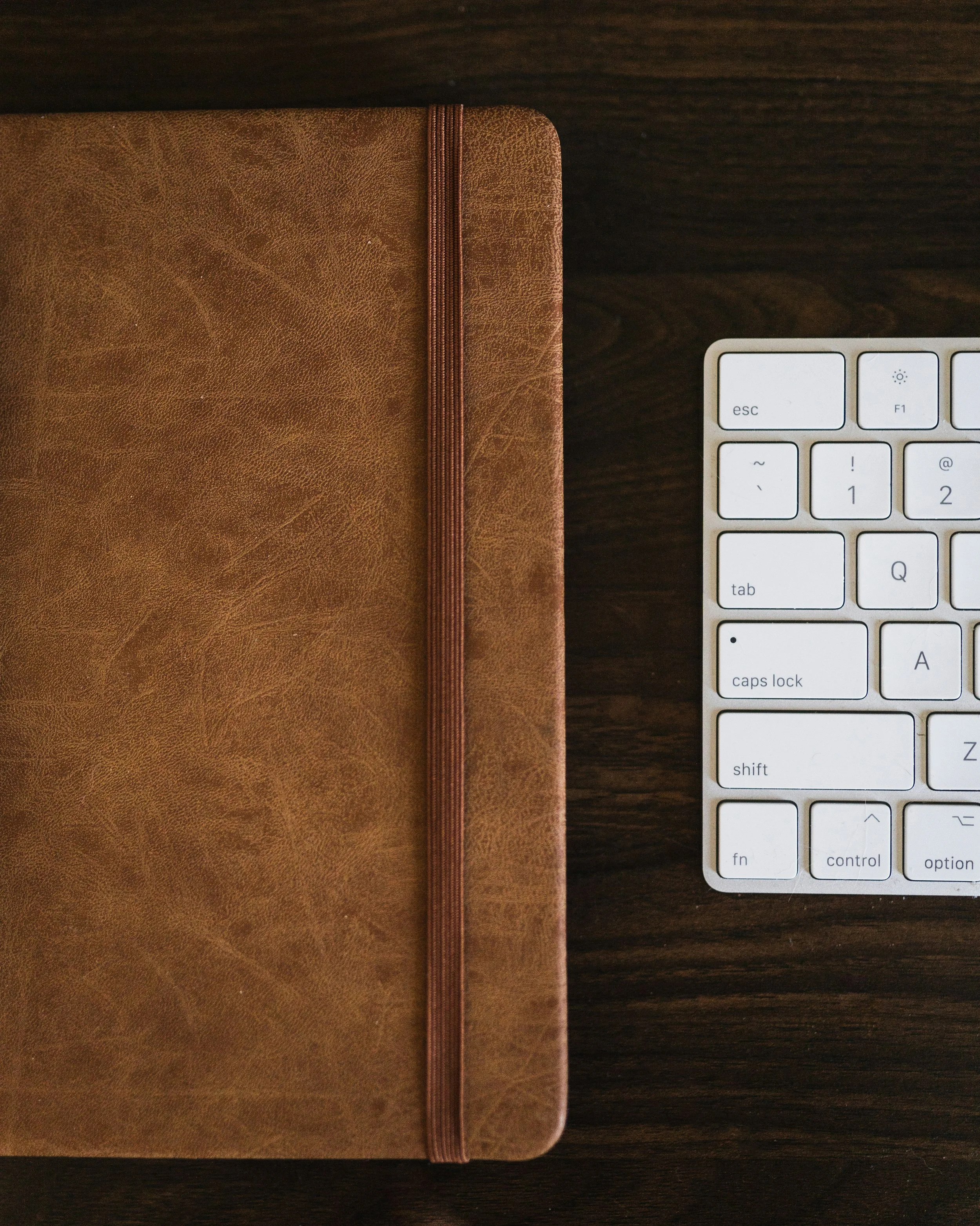Top view of a brown leather journal with a red elastic band next to a white keyboard on a dark wooden surface.