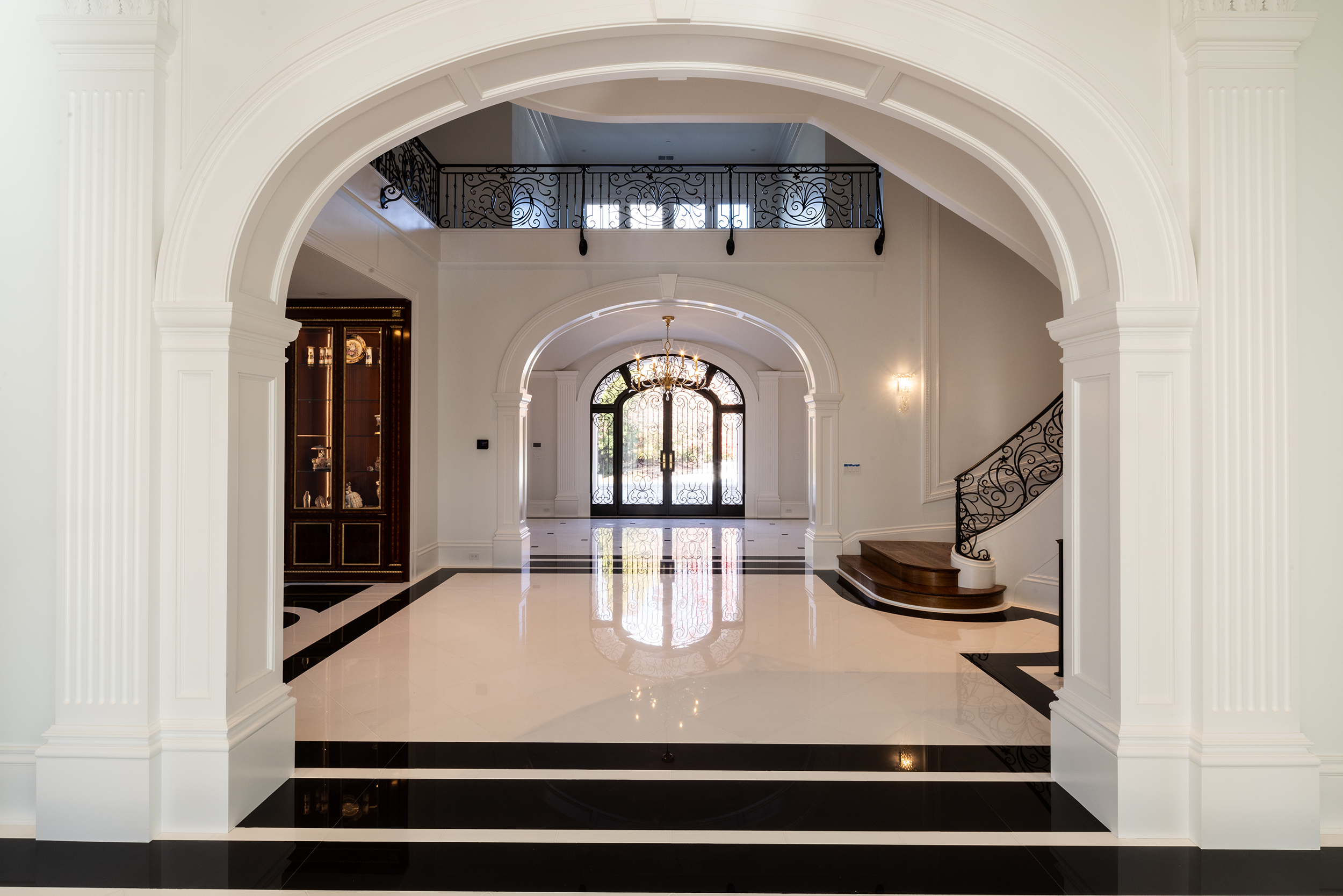 Elegant entryway with black and white checkered marble floor, white pillars, ornate black stairs, chandelier, black wrought iron door, and balcony railing.
